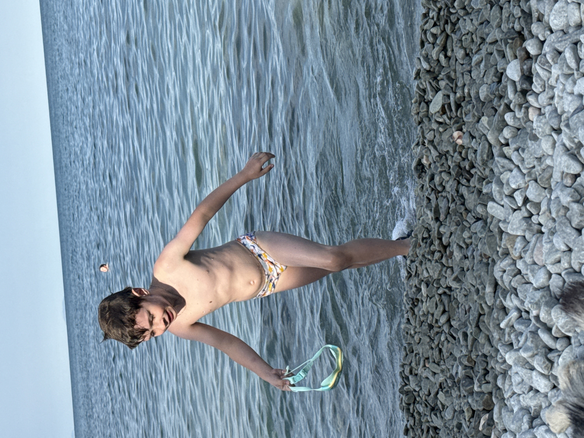 The image shows a young boy walking out of the water onto a rocky shore. He is wearing patterned swim trunks and holding a pair of swimming goggles. The boy appears to be in his pre-teen years. The water is calm with some gentle ripples, and the sky is clear and blue. In the background, another person can be seen in the water. The shore is covered in smooth, grey pebbles. The scene appears to be a natural, outdoor environment.