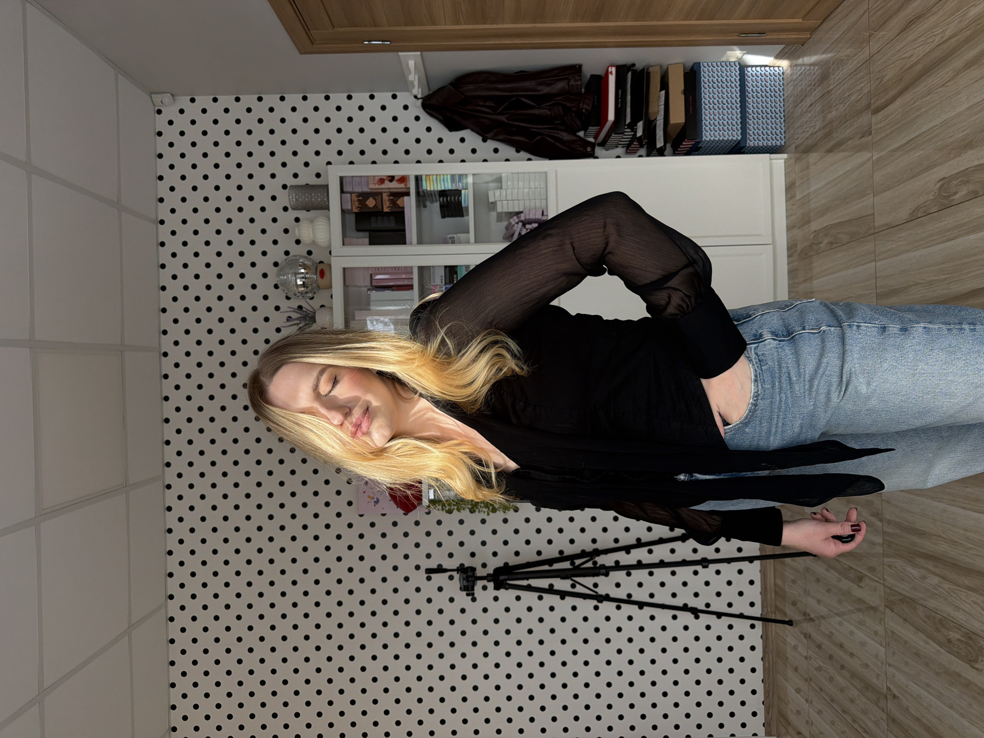 A woman with blonde hair lies on a wooden floor next to a wall with a black and white polka dot pattern. She is wearing blue jeans and a black shirt. In the background, there is a white cabinet with items on the shelves and in a cabinet above it. A black tripod is positioned near the woman. The location is unknown, and there is no evidence to suggest it is Riga, Latvia.