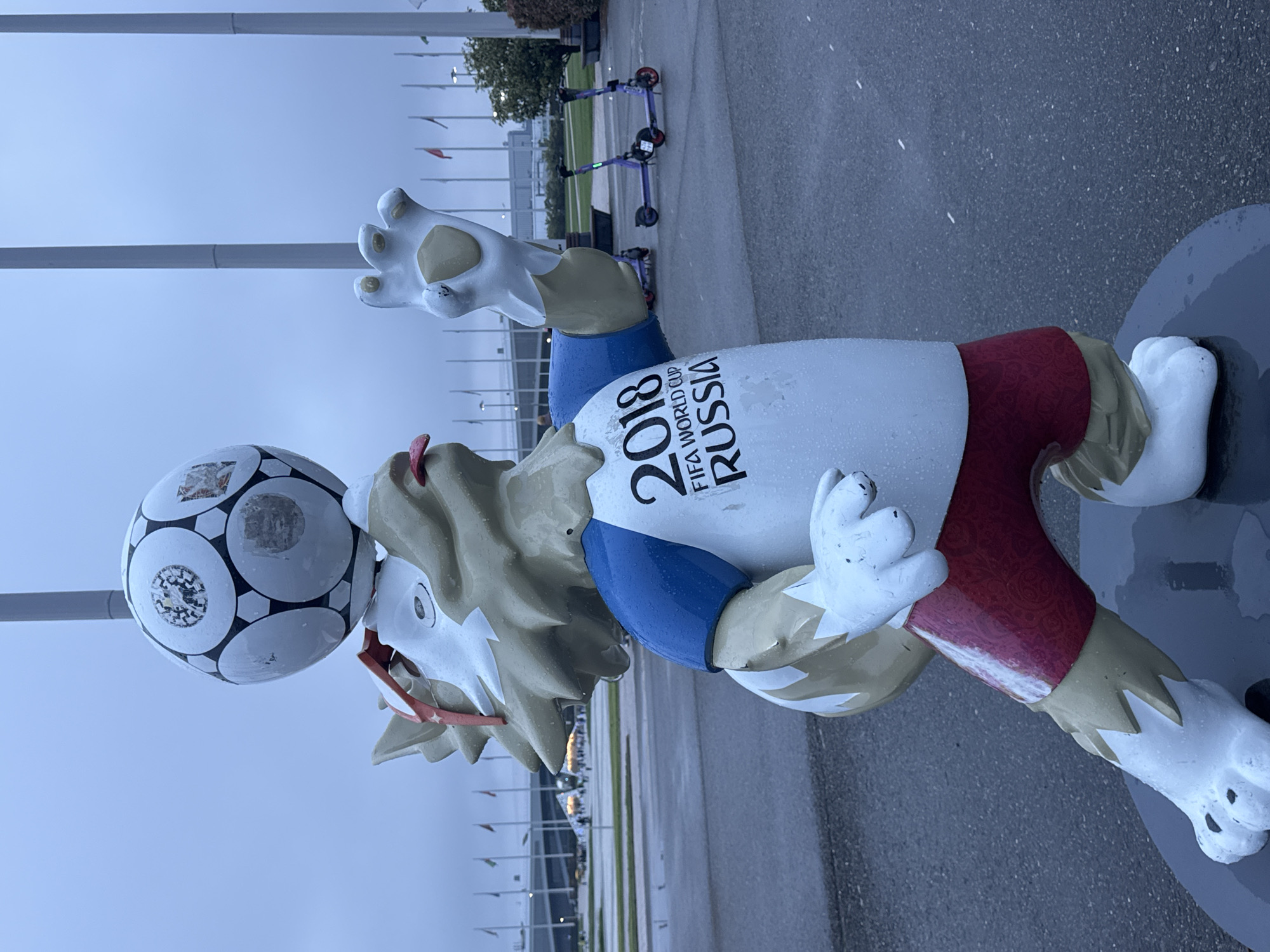 The image shows a statue of Zabivaka, the official mascot of the 2018 FIFA World Cup, held in Russia. The statue is located in Sirius, Russia. Zabivaka is depicted as a wolf wearing a white t-shirt with the words "2018 FIFA WORLD CUP RUSSIA" written on it, blue shorts, and red and white striped sneakers. The statue is standing on a grey pavement. In the background, there are flag poles, trees, and a few electric scooters. The sky appears overcast.