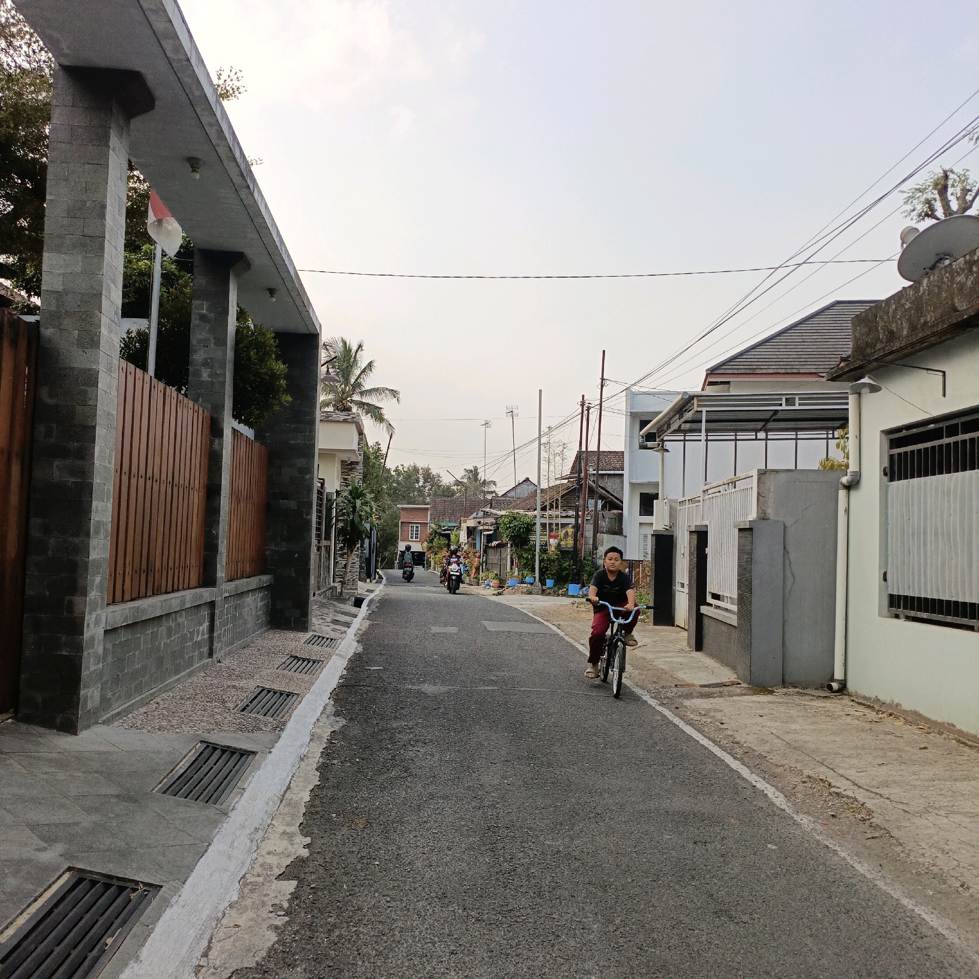 The image depicts a street scene in Salatiga, Indonesia, during daylight hours. In the foreground, a young person is riding a bicycle on an asphalt road. The cyclist wears a dark t-shirt and red pants. To the left of the road is a sidewalk with drainage grates. Beyond the sidewalk, a grey stone wall with a wooden fence runs along the property line, and a flag pole with an Indonesian flag is visible. Buildings line the right side of the street, some with visible satellite dishes and power lines overhead. In the background, other individuals on motorcycles are also present on the road. The sky is overcast.