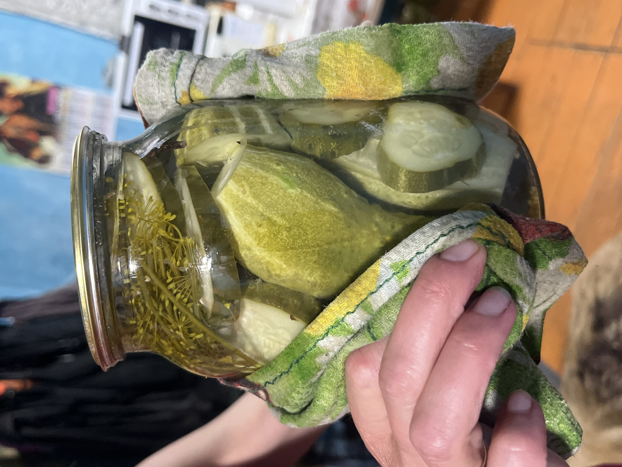 A close-up, slightly high-angle shot shows a glass jar filled with pickles and dill being held by a person's hand wrapped in a green and yellow patterned dishtowel. The jar is full of sliced cucumbers and dill sprigs, submerged in brine. The lid is a metallic silver color. The background is out of focus, showing a dark-colored garment on the left and a brightly lit wall with a poster and appliances on the right. The floor appears to be made of wood. The image does not provide any information about the city of Nizhny Tagil, Russia.