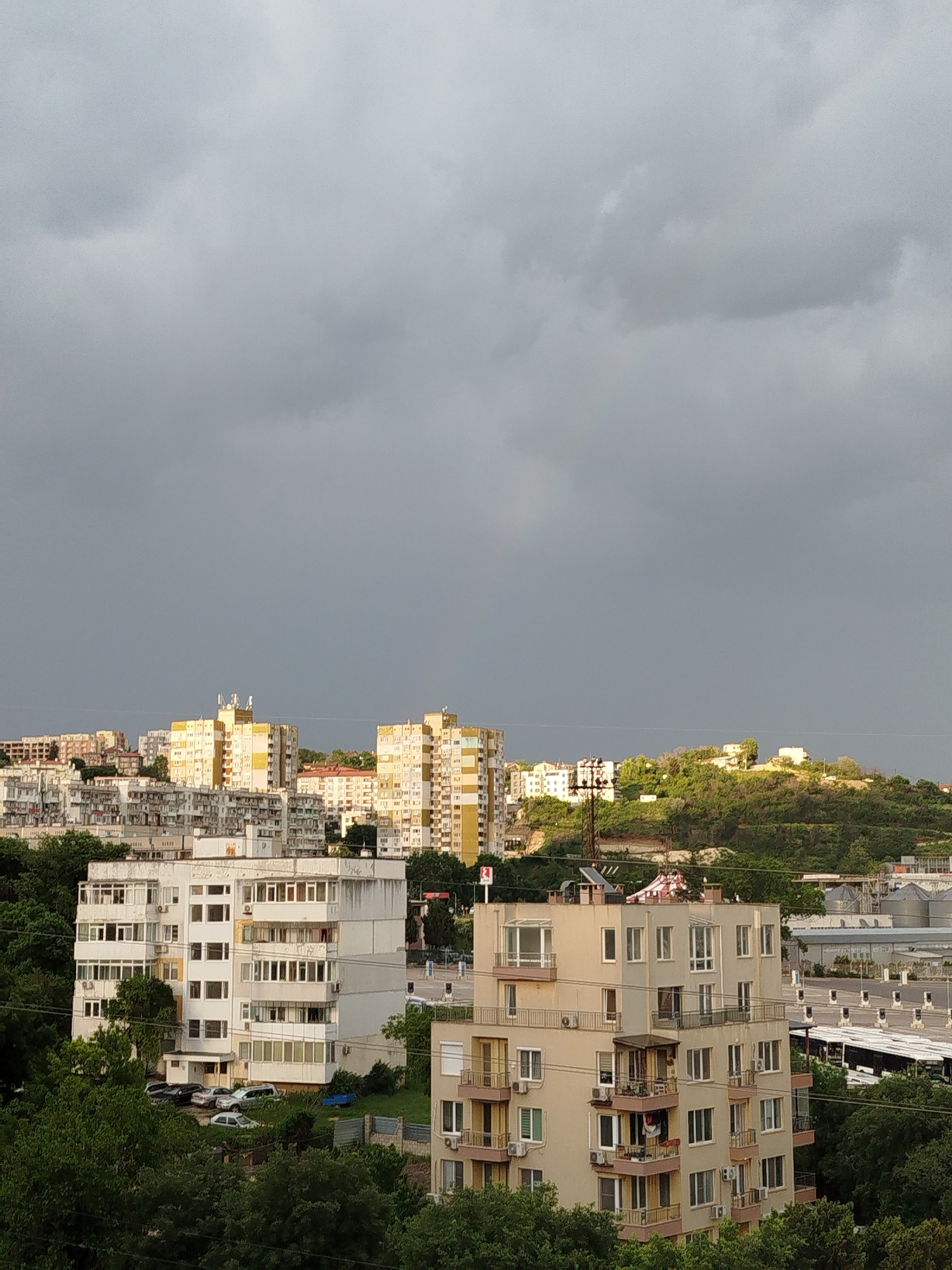 The image shows a cityscape in Varna, Bulgaria, under an overcast sky. In the foreground, there are two residential buildings, one white and one beige, surrounded by green trees. In the background, there are several taller, yellow and white apartment buildings. Further back, a green hill is visible with a few houses on top. To the right of the beige building, there are some industrial structures with silos. In the area between the two buildings and behind the white building, some parked cars are visible. The general mood is gloomy due to the gray sky.