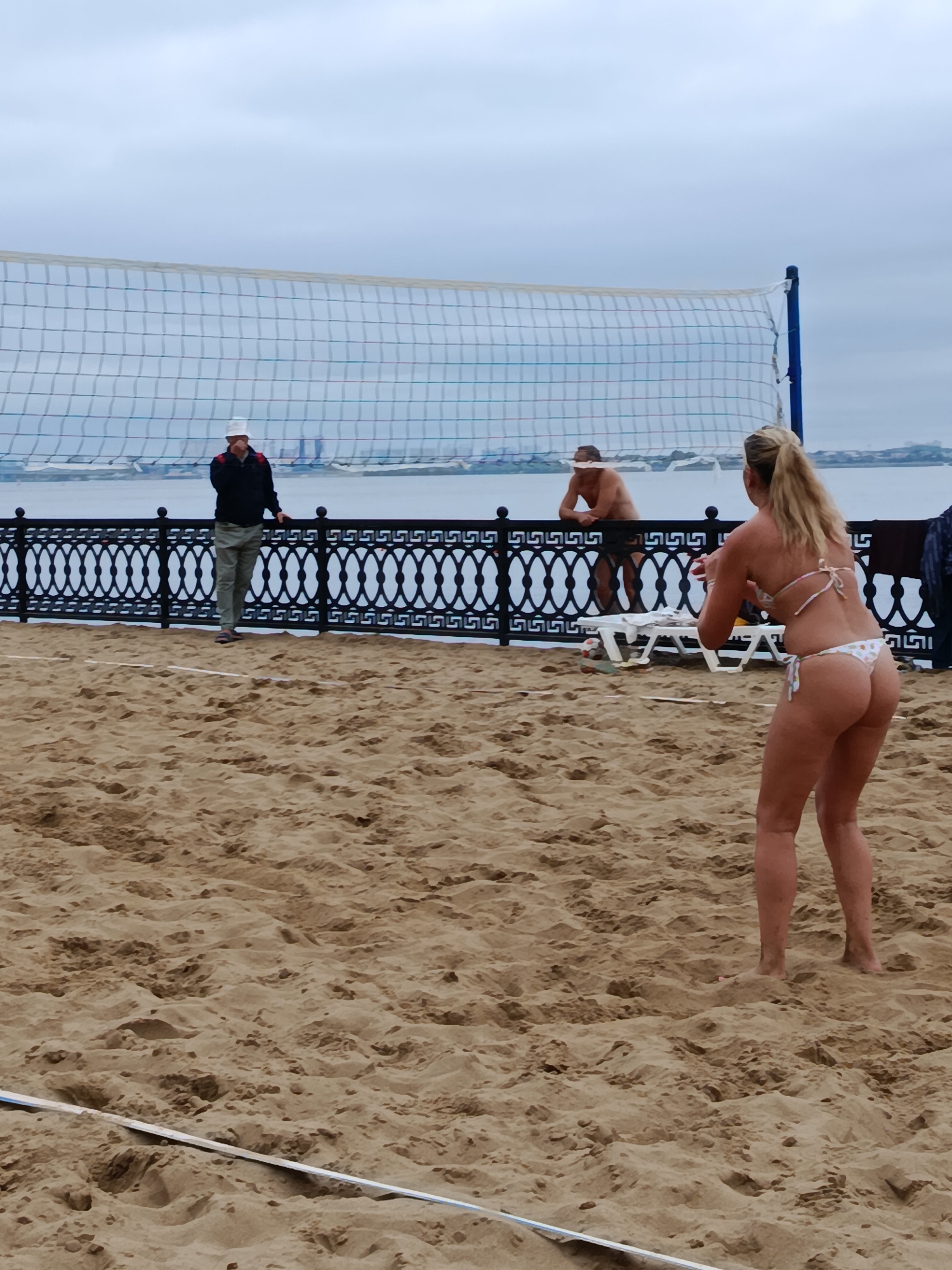 A group of people are playing volleyball on a sandy beach, with a net set up for the game. In the foreground, a woman in a bikini is poised to hit a volleyball. In the background, a man in a hat and a woman also in a bikini are on the other side of the net. A body of water can be seen beyond the beach, with buildings in the distance. The sky is overcast.