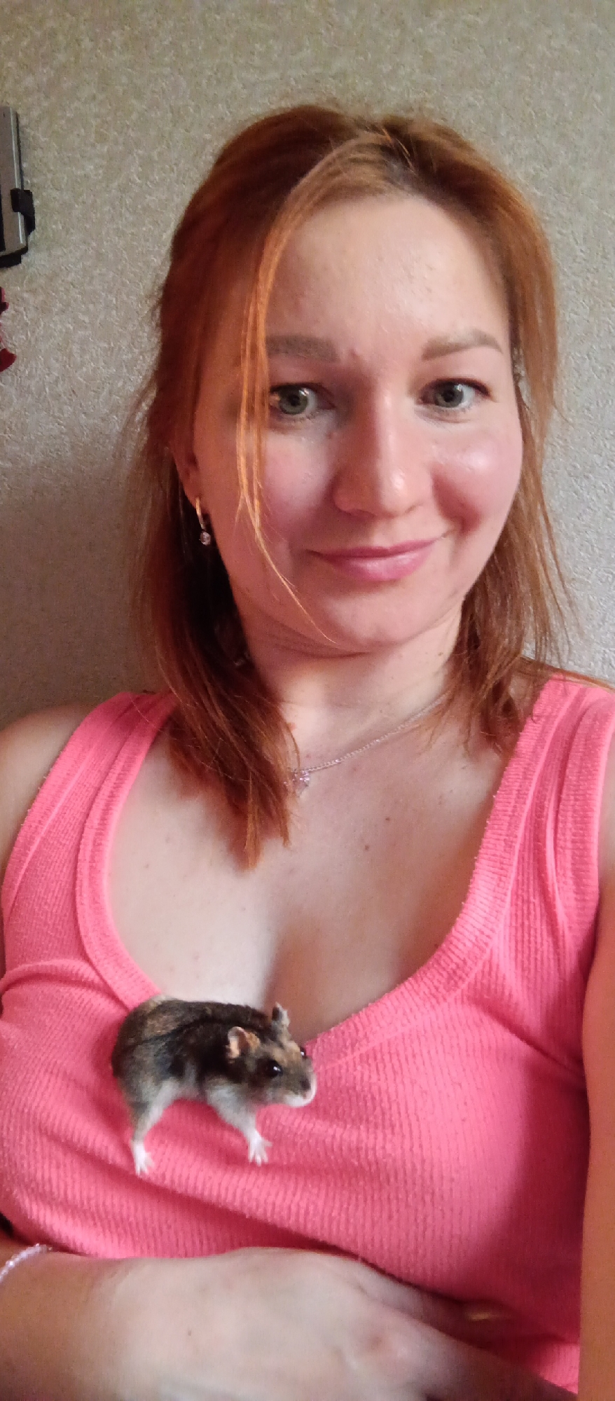 A woman with reddish-brown hair and fair skin is shown in a close-up shot wearing a pink ribbed tank top. A small hamster with dark brown and gray fur stands on the front of her shirt, near the neckline. The background is a textured, neutral-colored wall. The woman is smiling slightly, and she is wearing a silver necklace. She is also wearing silver hoop earrings. The photograph appears to be taken indoors with natural light.