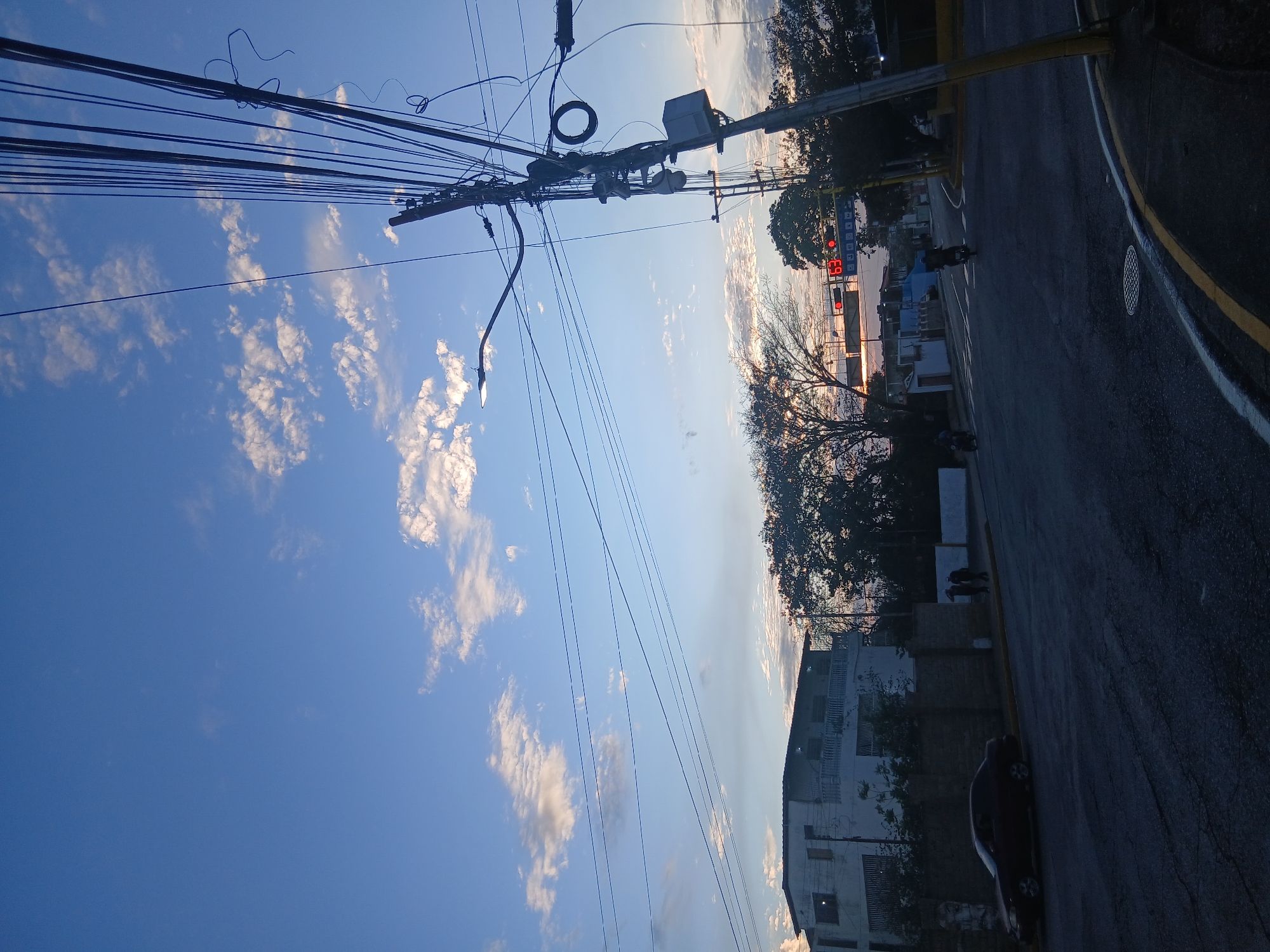 The image depicts a street in Juan Griego, Venezuela, captured at a rotated angle. Overhead power lines dominate the upper portion of the frame against a backdrop of a partly cloudy, blue sky. A utility pole is visible, supporting the power lines and a streetlight. On the right side of the frame, a street runs along with a yellow curb, a sidewalk, buildings, and trees. A traffic light displaying "69" can be seen. A motorcycle and a few pedestrians appear on the street, and a car is parked. The image is shot with a low sun angle casting long shadows.
