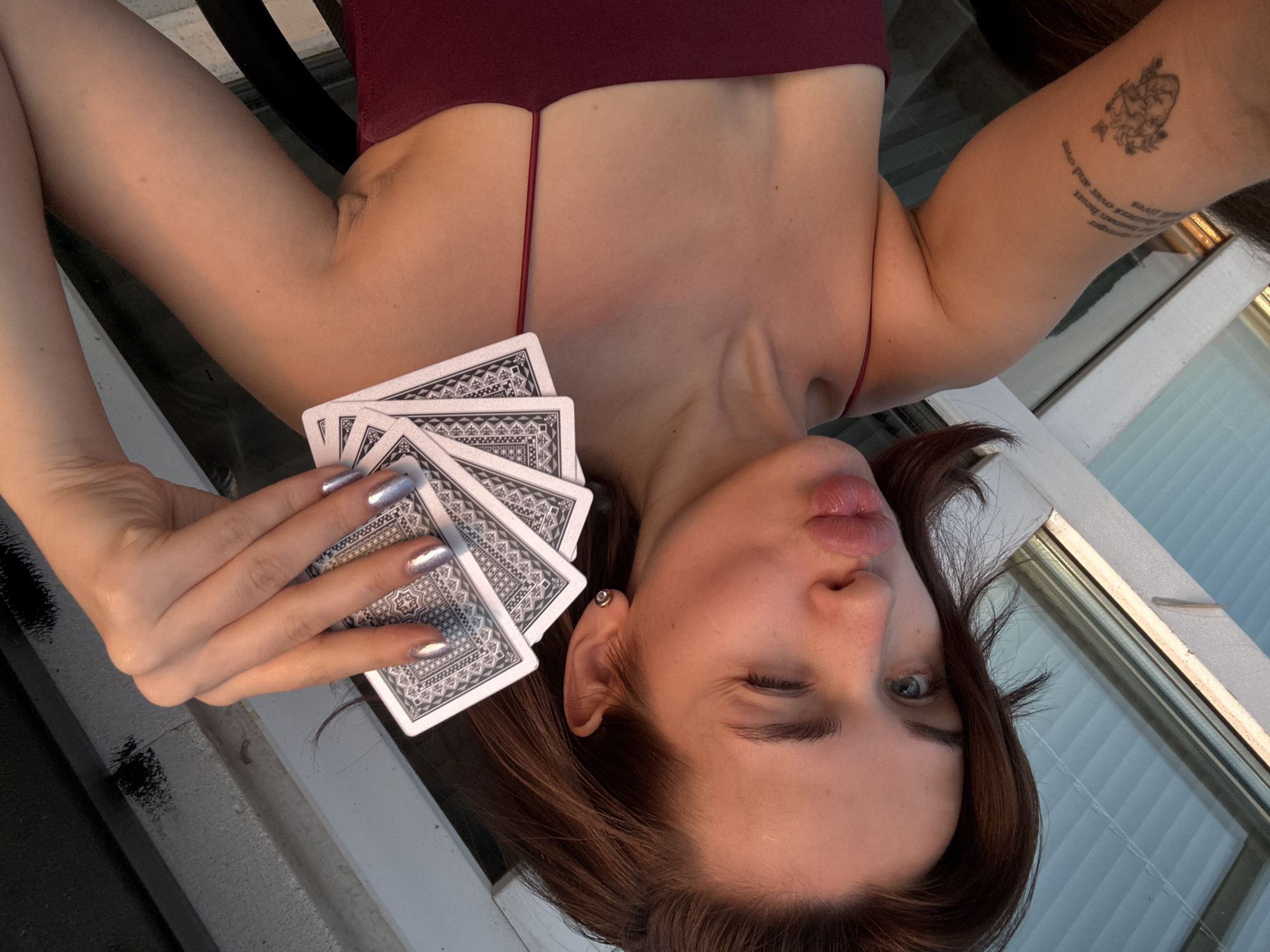 A young woman is lying on her back indoors, holding playing cards, winking, and puckering her lips. She is wearing a maroon tank top. Her nails are painted silver. She has a tattoo on her right arm. The background shows a window frame and a glimpse of blinds. There is no indication this image was taken in Лобаново, Russia.