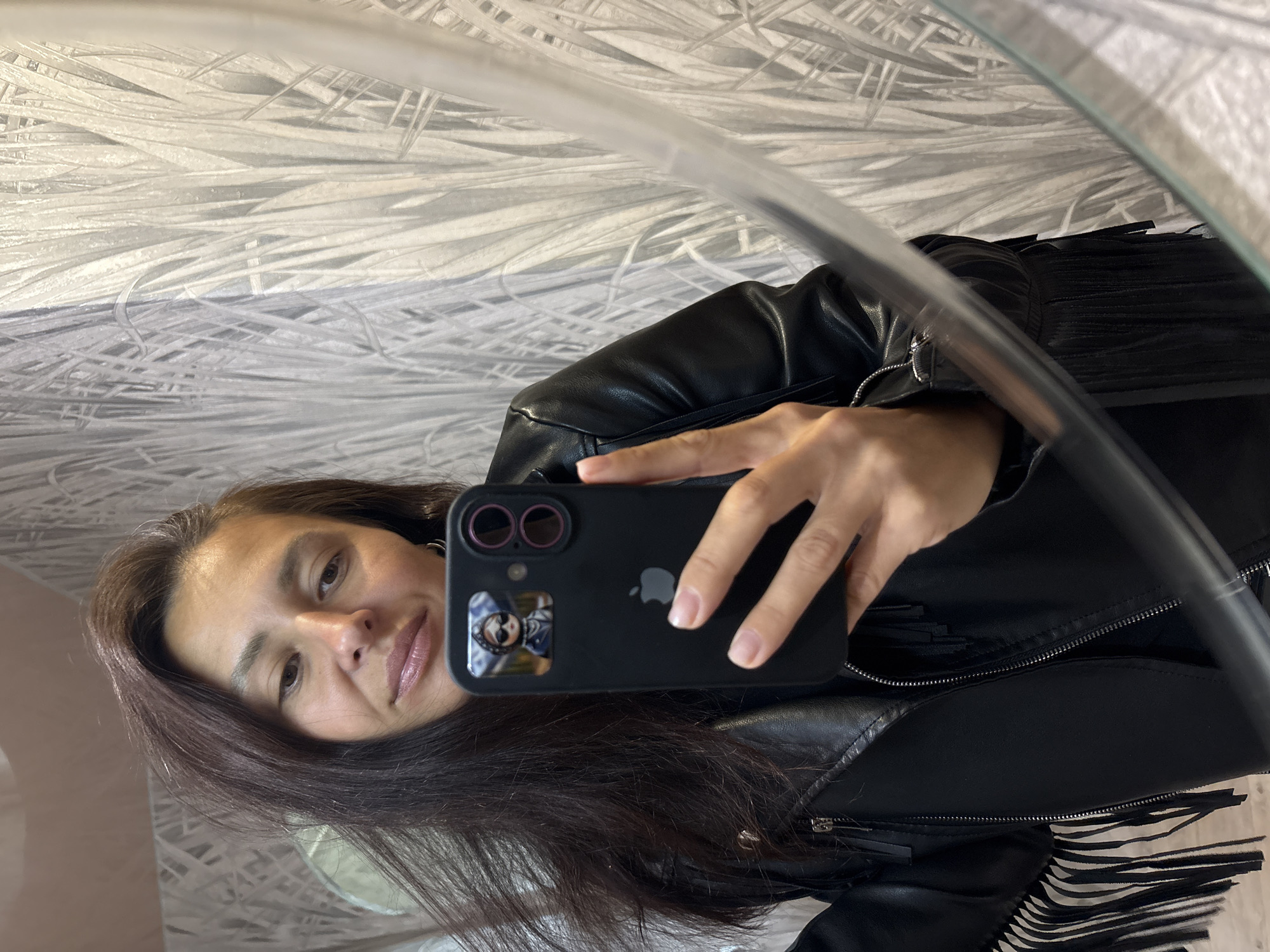 This image is a mirror selfie featuring a person with long, dark brown hair and light brown eyes. The person is looking directly into the smartphone they are holding, displaying a subtle, neutral smile.
The primary subject is holding a black iPhone in a black protective case, with the iconic Apple logo visible on the back. The phone case features two prominent camera lenses and a small, rectangular sticker or image of a person on its lower left side. The person's hand, holding the phone, has neatly manicured fingernails.
The individual is wearing a black leather jacket with distinctive fringe detailing visible on the sleeves and shoulders, suggesting a stylish or edgy aesthetic.
The setting appears to be indoors, reflected in a mirror. The background beyond the person reveals a light-colored, patterned wallpaper. The wallpaper has an abstract design of thin, interwoven, or layered lines and shapes, predominantly in shades of white, light gray, and subtle blue tones, resembling dried reeds or a woven texture.
There are no explicit cues for the time of day or weather. The only visible text is the Apple logo and the "iPhone" brand on the back of the phone. The general ambiance is calm and self-reflective. While the location context is Chaikovsky, Russia, no visual elements in the image directly indicate this geographical location.