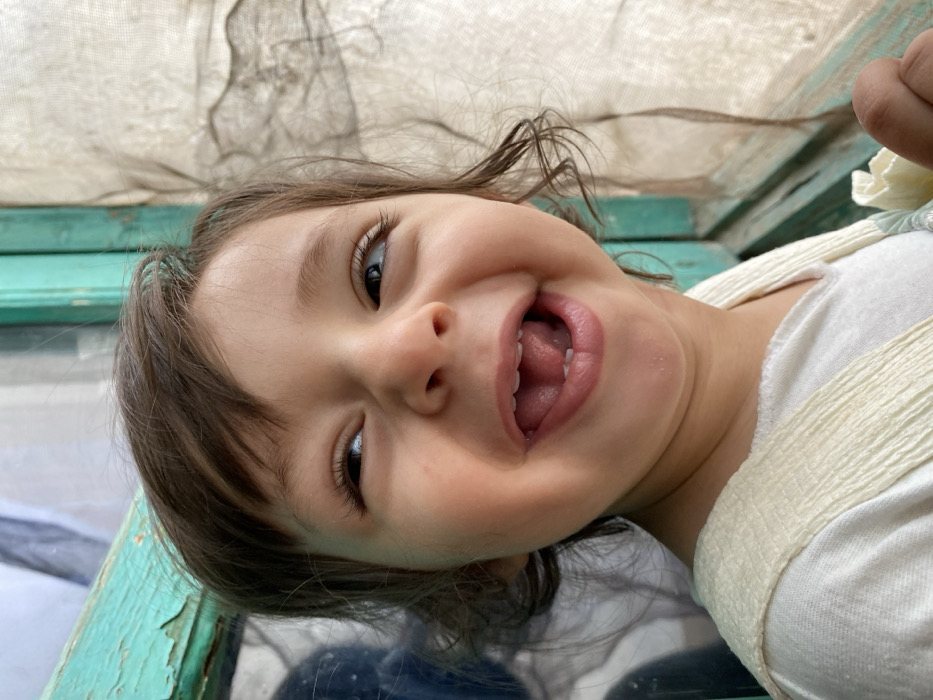 A close-up photograph depicts a young child against a background in Kandahar, Afghanistan. The child, with light brown hair and dark eyes, is captured in a wide, open-mouthed smile, with the tongue visible and lower front teeth exposed. The child's gaze is directed upwards and to the viewer's right. The child wears a light-colored shirt underneath a cream-colored, textured sleeveless garment. The background features a turquoise-painted wooden frame, likely part of a window or doorway, exhibiting peeling paint. To the right of the child, a translucent, light-colored woven fabric, resembling a screen or thin curtain, is visible. The child's right hand is partially observed in the bottom right corner, holding a light-colored, folded object.