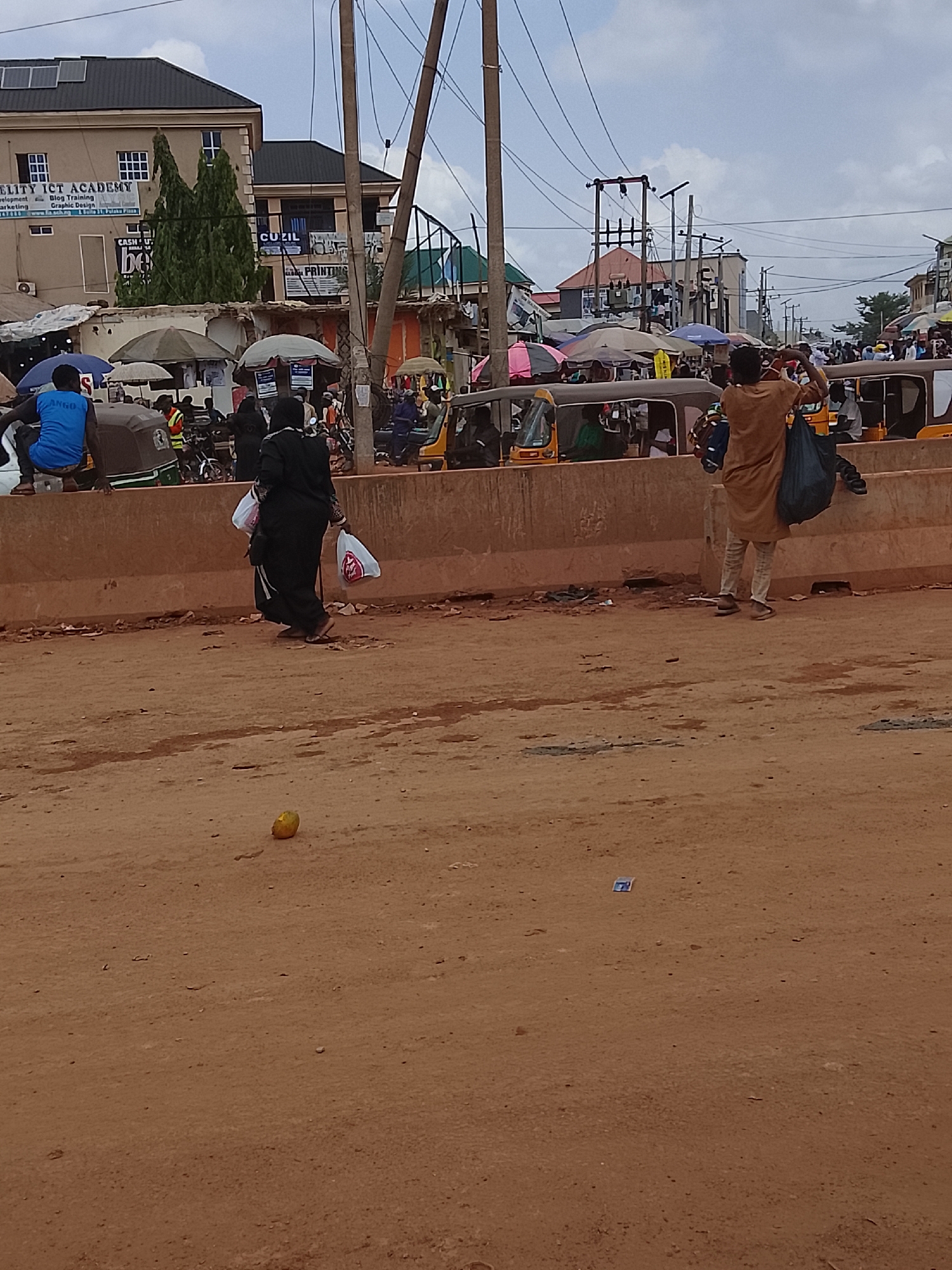 The image captures a street scene in Katambe, Nigeria. In the foreground, a dirt road is visible, with a yellow fruit and a small piece of paper littering the ground. A concrete barrier runs across the midground, behind which is a busy market street. People are standing and walking along the barrier. One person is wearing brown attire. Another figure, dressed in black, is seen walking across the street holding white shopping bags. The backdrop of the photo is a market scene with stalls covered by umbrellas, and three-wheeled vehicles and motorcycles parked near the stalls. Overhead are utility poles with a network of electrical wires. In the distance, buildings are visible. The sky is overcast.