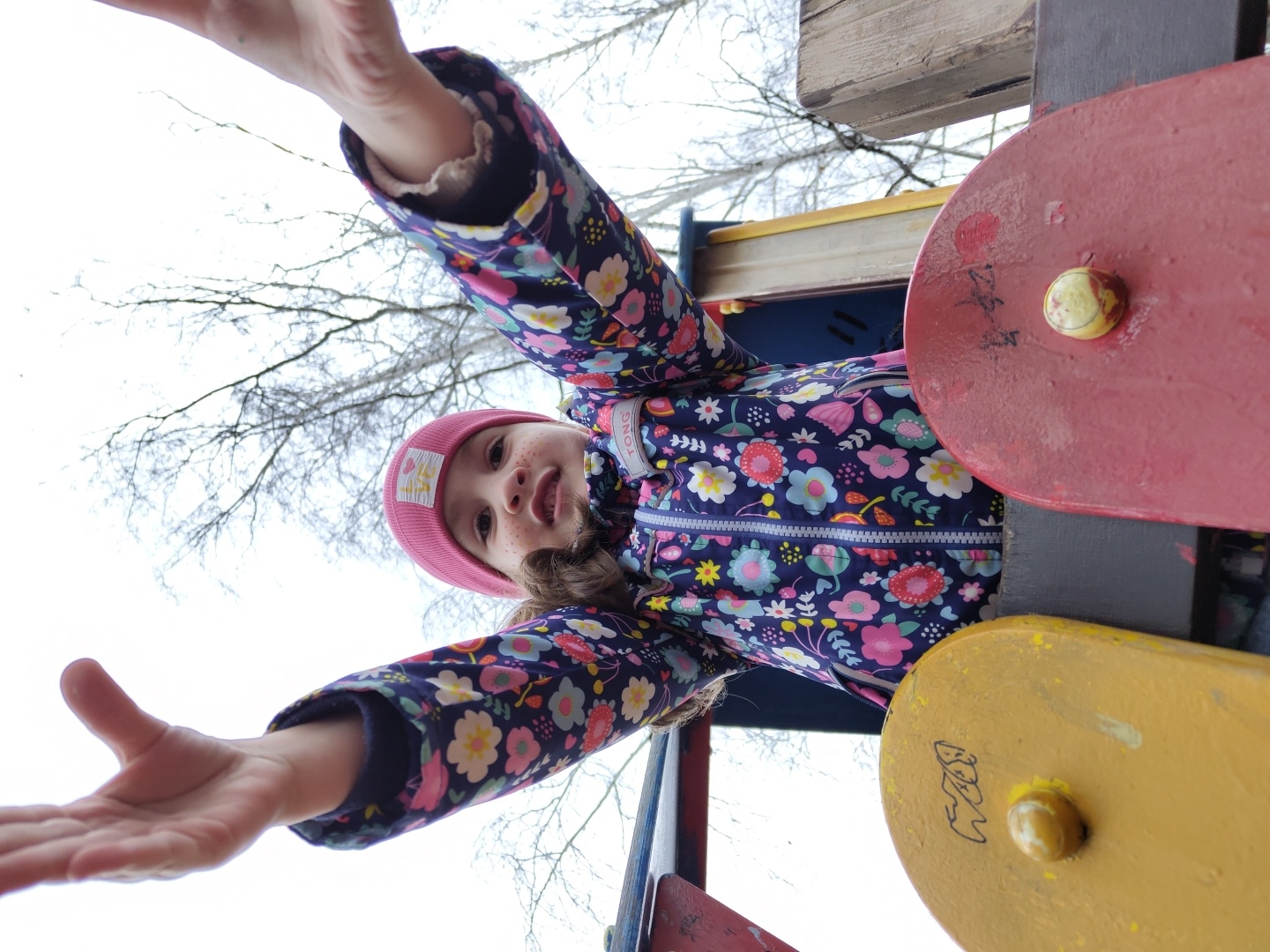 A low-angle shot captures a young girl at a playground in Chaikovsky, Russia. She wears a pink hat and a patterned floral jacket, extending her arms towards the camera. Playground equipment is visible, including red and yellow colored disks. Bare trees fill the background, suggesting a winter or early spring scene.