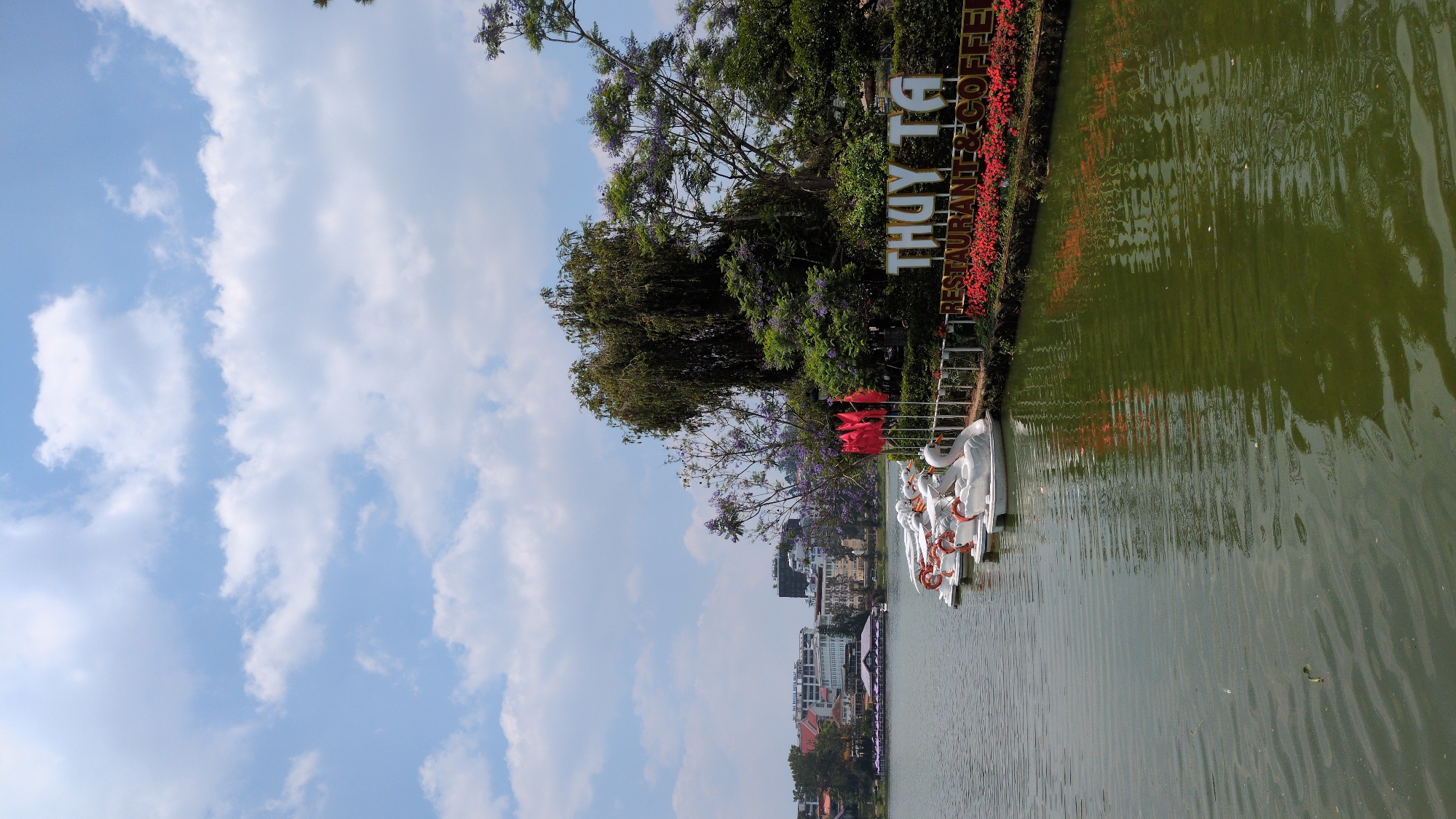 The image shows a lake in Da Lat, Vietnam, with swan-shaped paddle boats docked along the edge. A sign that reads “THUY TA RESTAURANT & COFFEE” is seen along the lakeshore. Red flags line the shore. Buildings are visible on the distant shoreline. The sky has scattered clouds.