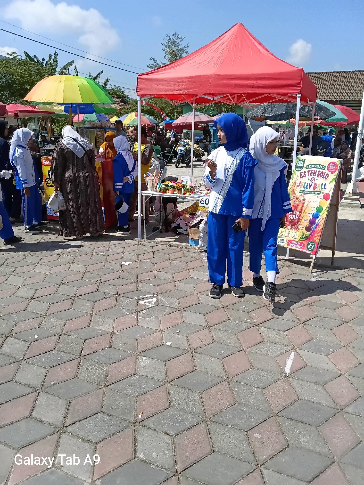 The image depicts an outdoor market scene in Ngadirenggo, Indonesia. In the foreground, two young women in blue and white uniforms, likely school uniforms, stand on a patterned brick pavement. One woman holds a mobile phone. Behind them, a red tent covers a market stall. Several other market stalls with colorful umbrellas are visible in the background, along with people browsing and interacting. A sign promoting "ES TEH SOLO & JELLY BALL" with vibrant imagery is prominently displayed near the two women. The environment is sunny with a bright blue sky.