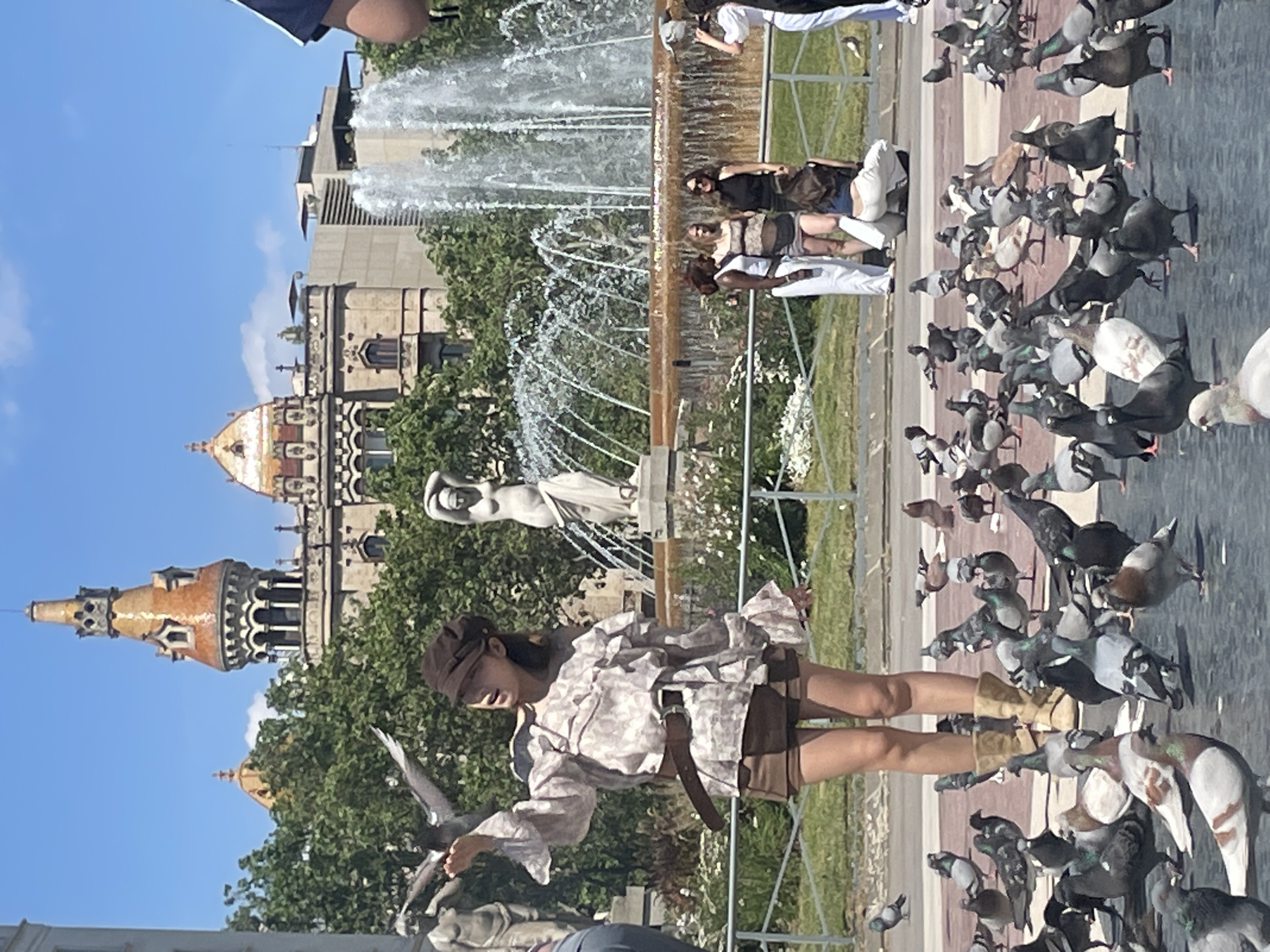 This media file captures a vibrant, sunny daytime scene in a public park or plaza in Barcelona, Spain, likely the Parc de la Ciutadella, given the distinctive architecture and grand fountain.
In the foreground, a person wearing a light-colored, patterned long-sleeved shirt, brown shorts, and a brown cap stands on a paved area, actively interacting with a flock of pigeons. One pigeon is perched on their outstretched hand, while numerous others are gathered around their feet and scattered across the ground, suggesting they might be feeding the birds.
The midground is dominated by a majestic, multi-tiered fountain (Cascada Monumental) with water vigorously spraying from multiple jets. Near the fountain, several other individuals are visible, observing the surroundings. A classical white statue of a figure stands prominently within the fountain's design.
In the background, under a clear blue sky, distinctive architectural structures are visible. These include an ornate building with intricate detailing and a prominent tower topped with an orange-tiled, pointed roof and a smaller dome, characteristic of the Castell dels Tres Dragons within the park. Lush green trees and grass flank the paved areas and the fountain, adding to the park-like setting. The weather is bright and clear, indicating a pleasant day. There is no visible text.