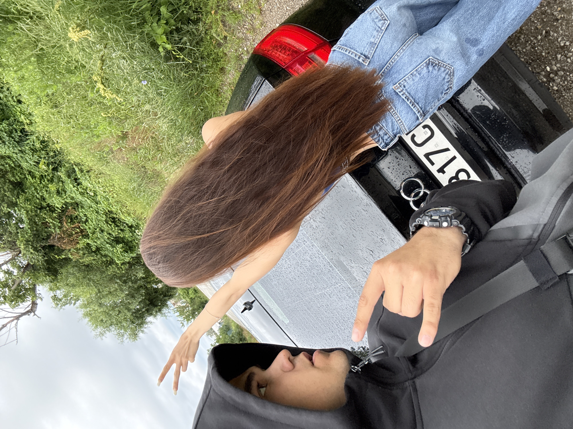 The image captures a close-up, selfie-style view of a young man and woman posing in front of a black car. The man, on the left, is wearing a black hooded jacket and looking towards the camera with a surprised expression. His right hand is raised, displaying a rock-on hand gesture. The woman, on the right, has long, dark brown hair and is leaning against the car. Her back is to the camera, and her left arm is extended, with her hand forming a peace sign. She is wearing blue denim jeans and a blue top, the latter partially visible. The car, a black Audi, is wet, indicating recent rain, with water droplets visible on its surface. The license plate, partially obscured, reads "B17C". The background consists of lush green vegetation and trees, with a cloudy sky overhead. The scene appears to be outdoors, possibly by a roadside or in a rural area.