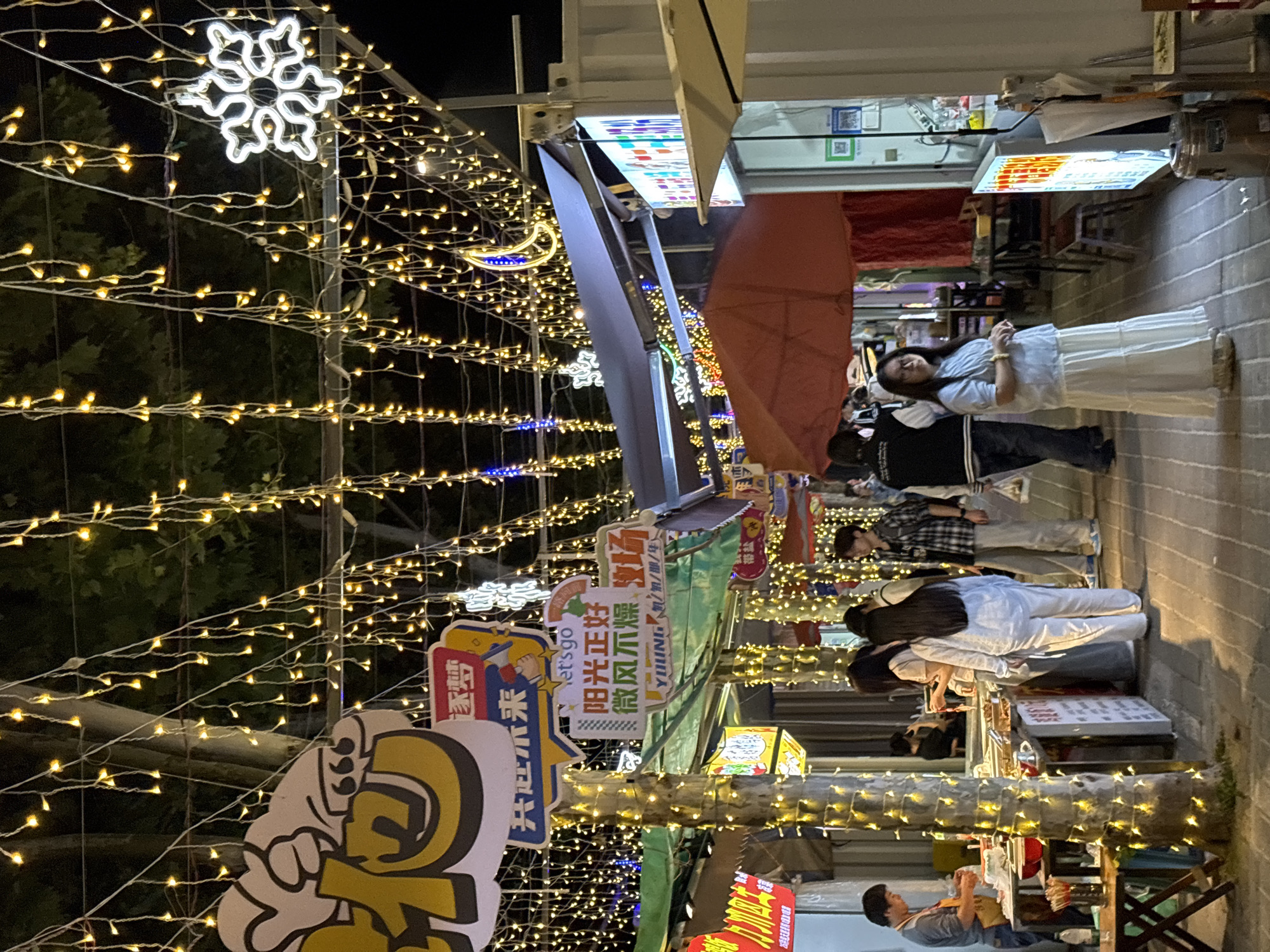 The image depicts a night scene on a street in Jiangjin, China, adorned with numerous strings of small, warm-toned lights. A large decorative snowflake is visible among the lights. Several people are present, including some browsing stalls and others simply walking through the area. Signs in Chinese are visible, indicating local businesses or promotions. A red umbrella is partially open over one of the areas where people are browsing. The setting suggests a bustling, lively atmosphere, possibly a night market or festive gathering.