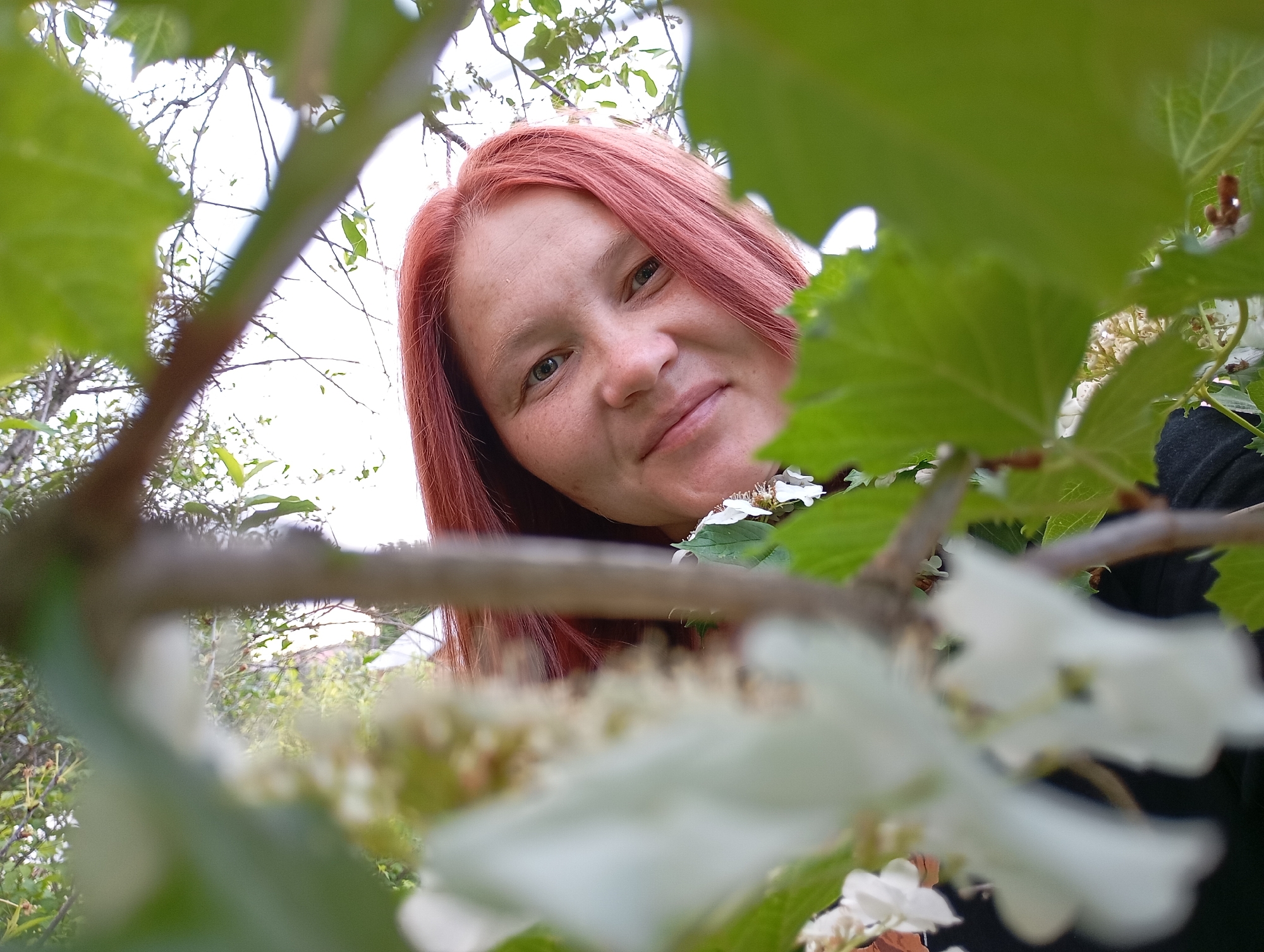 A woman with red hair looks down at the camera through green leaves and white flowers. The location is likely outdoors in a garden or natural setting in the city of Фоки, Russia.
