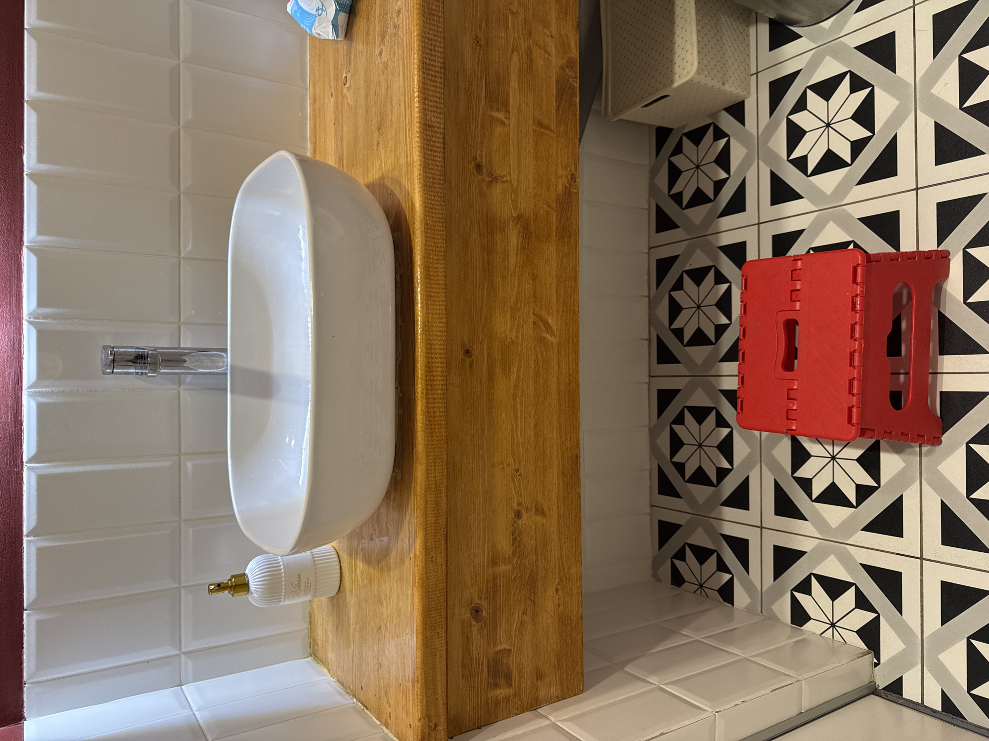 This media file captures a detail of a bathroom or lavatory space in a home in Patrusheva, Russia. The scene features a white urinal mounted on a wooden column. The wall behind the urinal is tiled with white subway tiles. To the right of the urinal, there is a section of floor tiled with a black and white geometric pattern, and a red foldable stool is visible. A white soap dispenser with a gold pump is attached to the wall near the base of the urinal. The lighting suggests it is daytime, likely indoors. No people are visible. There is some text on the soap dispenser, "Ditta ssa", and on a visible item at the top of the image, "Tork".
