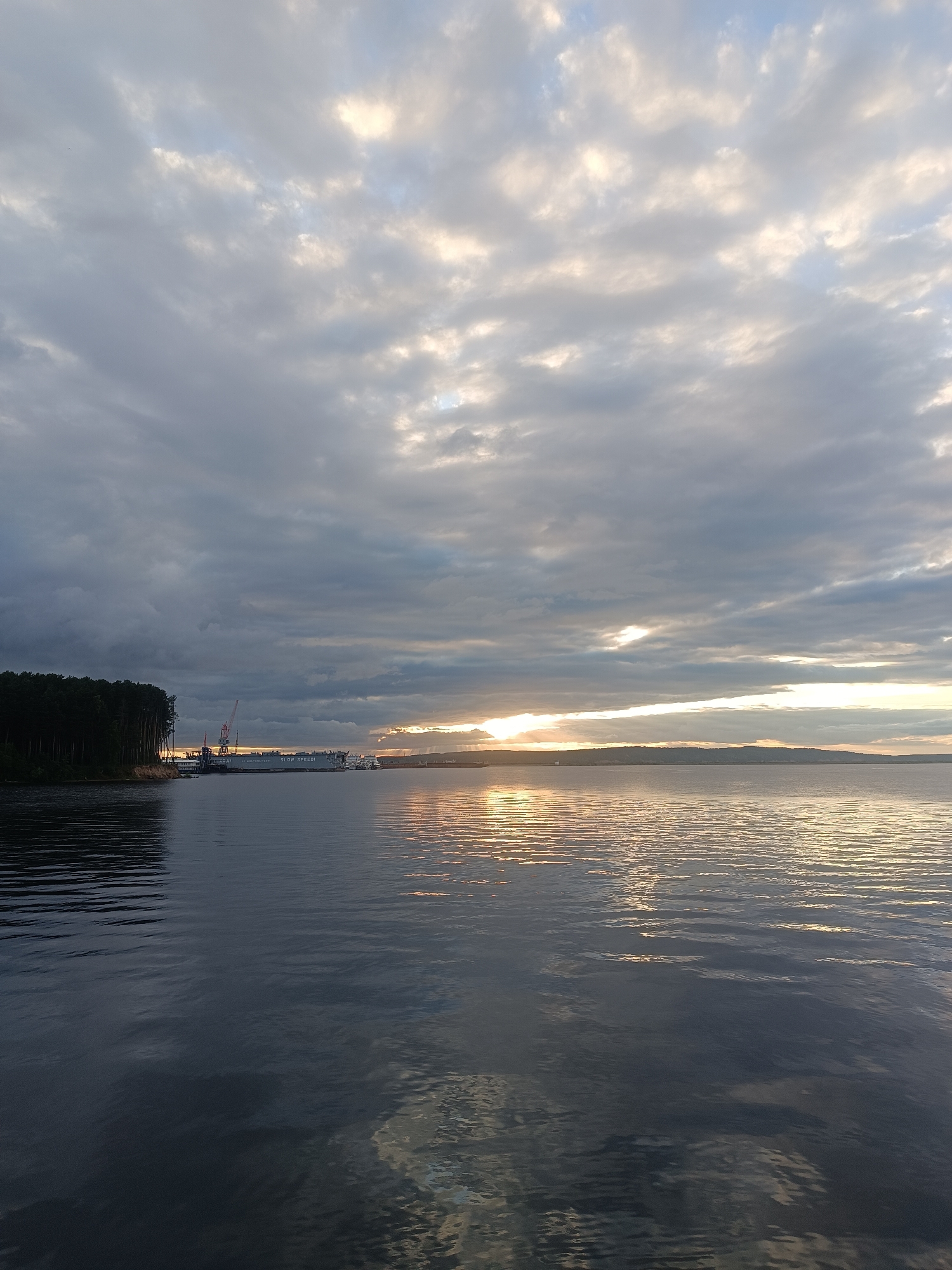 The image captures a serene sunset over the Kama River in Chaikovsky, Russia. On the left bank, a dense forest of dark green trees stands tall, meeting the calm, dark water of the river. Along the horizon, a port facility is visible, featuring cranes and several ships, one with "SLOW SPEED" printed on its side. The sky is a dramatic canvas of heavy, gray clouds, with intermittent breaks allowing the golden hues of the setting sun to pierce through, casting shimmering reflections on the rippling water. The overall atmosphere is peaceful, with the natural beauty of the landscape contrasting with the industrial presence of the port.