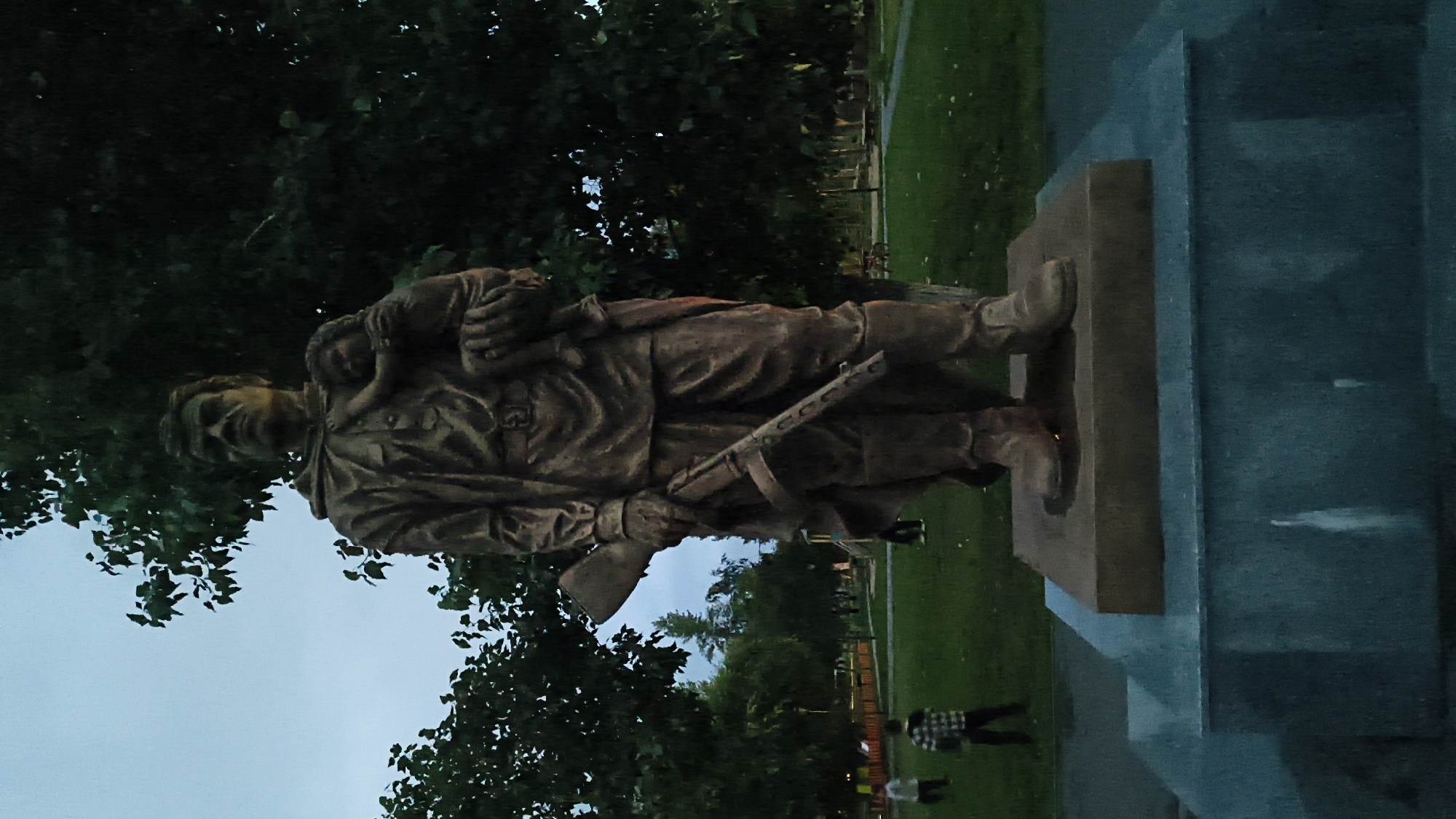 The image shows a bronze statue of a soldier holding a child and a rifle, located in Mikhaylovka, Russia. The statue stands on a multi-tiered stone base in a park setting. The soldier is depicted wearing military attire, including a hooded cloak and boots, and holds a submachine gun in his right hand. The child is cradled in the soldier's left arm. Behind the statue, trees and a lawn are visible. In the background, several people, including children, are present in the park. The sky is overcast, indicating a twilight or cloudy day. The overall scene suggests a memorial or monument dedicated to a soldier and child, likely commemorating a historical event or figure significant to the region.
