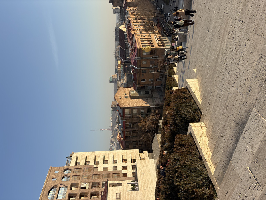 The image depicts the Yerevan Cascade in Yerevan, Armenia, during the late afternoon under a clear blue sky. The foreground features wide, light-colored stone steps descending from the viewer's perspective. To the left of the steps, terraced planters are filled with dense, dark green and brownish bushes. Several individuals are partially visible among these bushes.
On the main staircase, a group of approximately 10-15 individuals is walking downwards. They are casually dressed in various outerwear, with some carrying backpacks or bags. Their faces are largely indistinct due to distance and lighting.
To the left, a tall, modern building with light-colored stone facades and numerous rectangular and arched windows rises prominently. Beyond the bushes and the modern building, the urban landscape of Yerevan unfolds. Older, multi-story buildings constructed from reddish-brown tuff stone, featuring balconies and red-tiled roofs, dominate the midground. One specific older building on the right side of the midground displays a large, bright yellow, stylized humanoid figure attached to its exterior corner.
Further in the background, more city buildings of varying heights extend towards the horizon. The distinct Yerevan TV Tower is visible on a distant hill to the left of the center, and additional hills frame the cityscape in the distance. A street with vehicles is visible in the lower right, bordering the older buildings. The scene is illuminated by low-angle sunlight, casting long shadows.