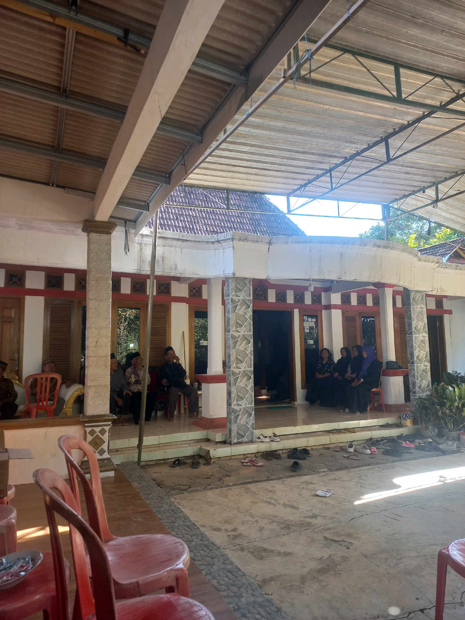 The image depicts an outdoor gathering in front of a traditional Indonesian house in Sukoharjo, Indonesia. The house features a white facade with red and brown decorative trim and wooden shutters on the windows. A covered patio area extends from the house, supported by columns.
Several individuals are seated on a raised platform in front of the house. On the left side, a group of men, some wearing traditional Indonesian attire like batik shirts and peci hats, are seated on chairs or a couch. On the right side, a group of women, all wearing hijabs or headscarves and dark clothing, are also seated.
The ground in front of the patio is paved with concrete and has a patterned edge made of small stones and larger grey stones. Numerous pairs of sandals and shoes are scattered on the ground near the entrance, indicating that attendees have removed their footwear before entering the house or gathering area. Some red plastic chairs are placed in the foreground. In the background, trees and foliage are visible, suggesting a rural or semi-rural setting. The sunlight casts distinct shadows on the ground, indicating it is daytime.