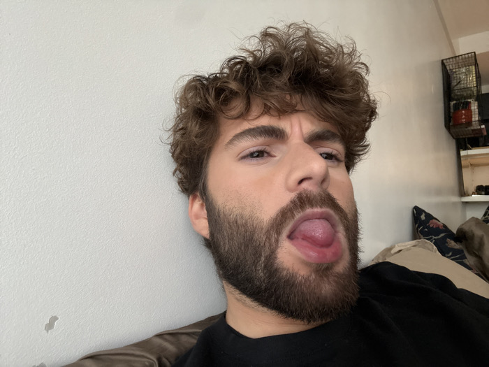 The image depicts a close-up, head-and-shoulders shot of an adult male in an indoor setting. The individual has fair skin, dark brown curly hair, and a full dark brown beard and mustache. He is wearing a black crew-neck garment. His mouth is open, and his tongue is extended outwards. His eyebrows are furrowed, and his eyes are directed towards the camera.
The background on the left consists of a plain, textured, off-white wall, with a small, irregular dark mark visible on its lower portion. On the right, a dark metal grid-like shelf or storage unit is mounted on the wall. Below the shelf, a portion of a dark-patterned cushion or pillow is visible, resting on what appears to be a brown upholstered furniture piece. The location within the city of Paris, France, cannot be determined from the visual information present in the image.
