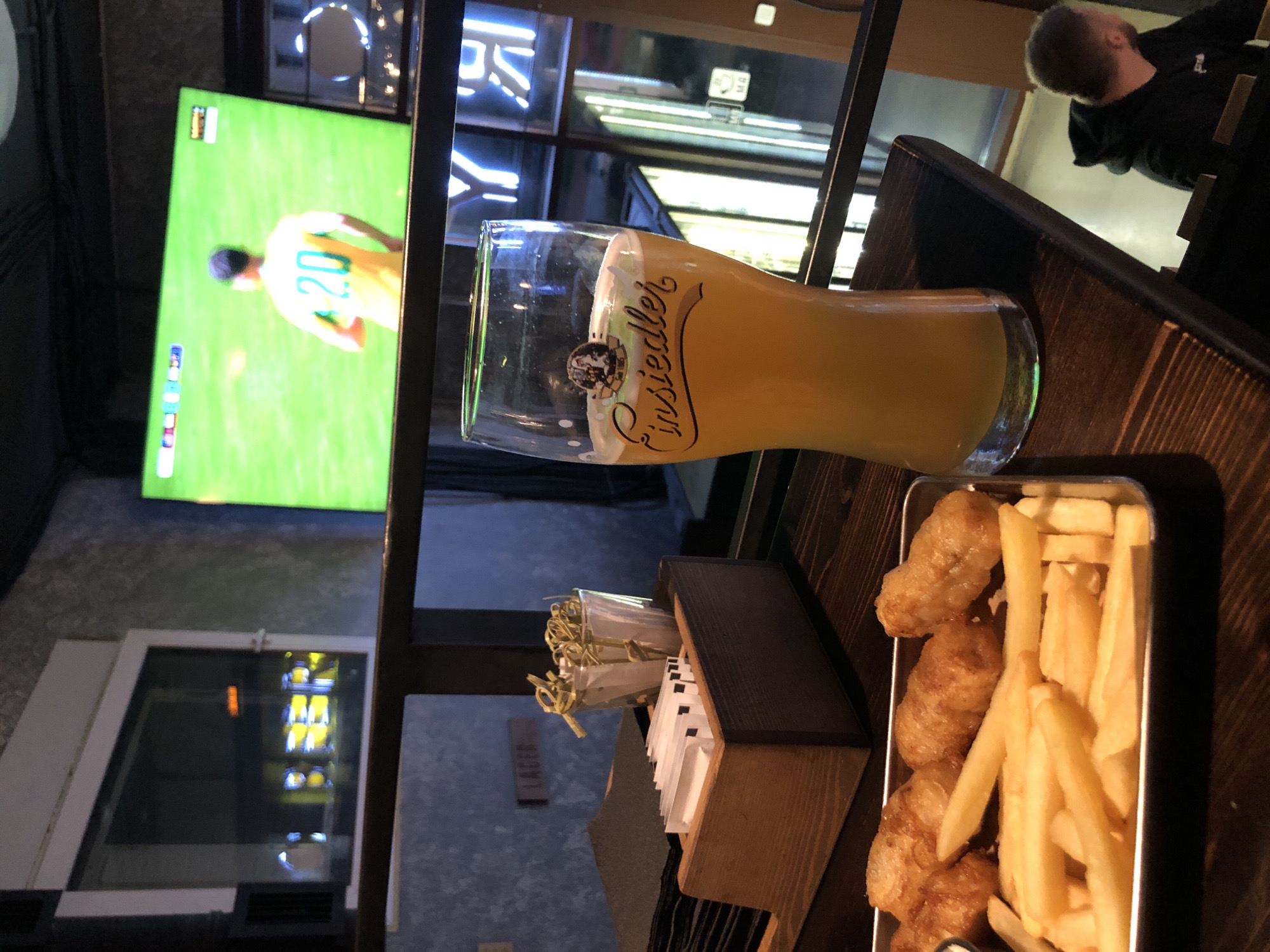 The image shows a close-up of a glass of beer and a tray of food, likely fish and chips, on a wooden table in what appears to be a bar or restaurant. In the background, a large television screen displays a soccer match, with a player wearing a yellow jersey with the number 20 visible. There is also a neon sign with Cyrillic lettering and a sign that reads "LAGER". A person is visible in the lower right corner, looking away from the camera. The overall setting suggests a casual atmosphere, possibly a sports bar in Saint Petersburg, Russia, given the presence of Cyrillic lettering and the implied casual dining experience.