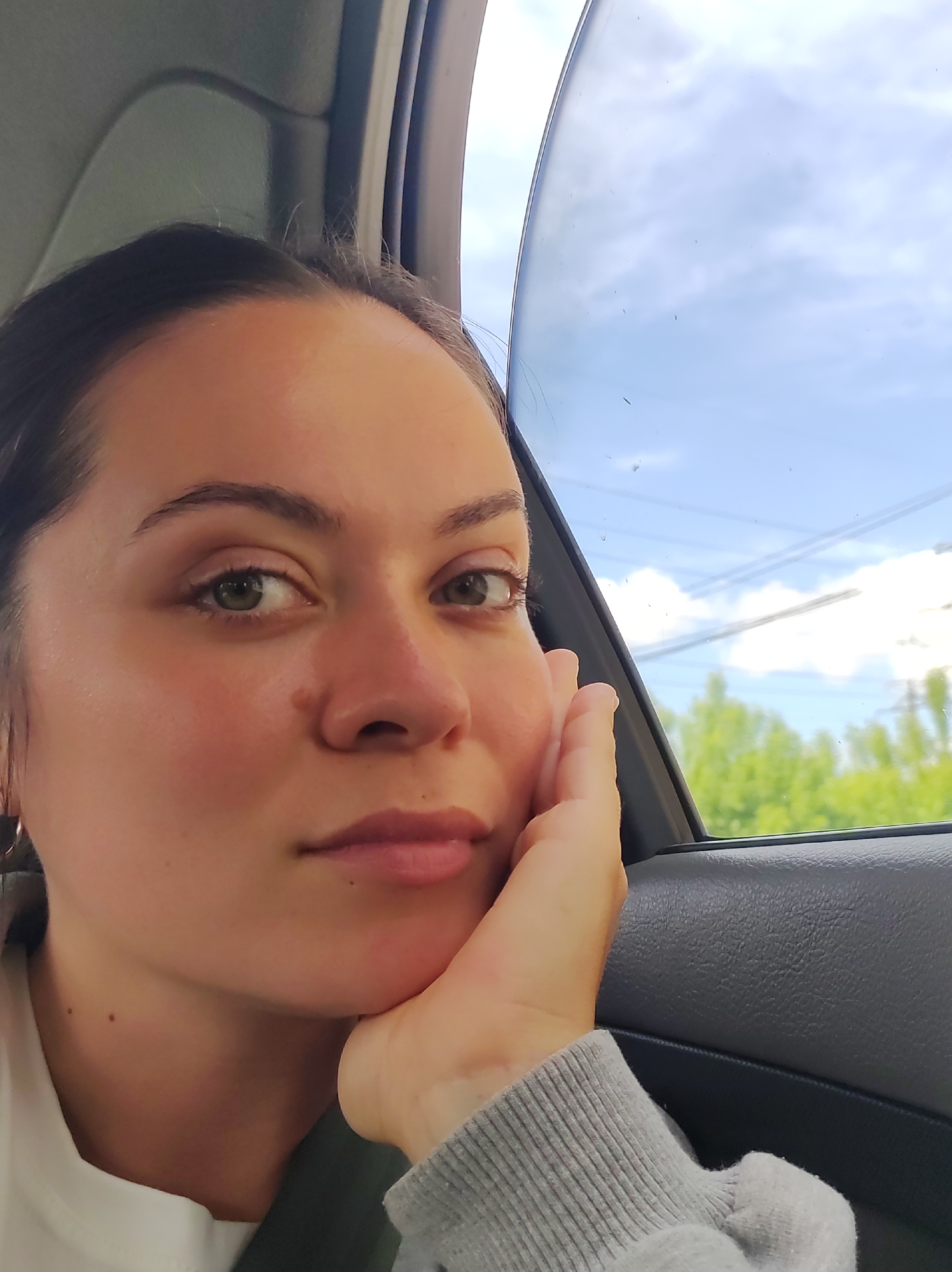 A close-up portrait shows a young adult female from the chest up, seated inside a car, likely in Chaikovsky, Russia. She is positioned towards the camera, with her head slightly tilted and her left hand gently supporting her jaw. Her expression is pensive and thoughtful, with direct eye contact. She has dark hair pulled back, green or hazel eyes, and appears to be wearing a light-colored top under a grey sweatshirt. A seatbelt is visible across her left shoulder.
The background shows the interior of the car, including a dark grey door panel and window frame. Through the car window, a bright daytime sky with scattered white clouds is visible. Lush green trees line the horizon, suggesting an outdoor scene in a rural or suburban environment. Power lines are visible in the distance against the sky and greenery. Small specks, possibly dust or light water marks, are present on the car window. The weather appears fair, with bright natural light illuminating her face. No visible text is present in the image.