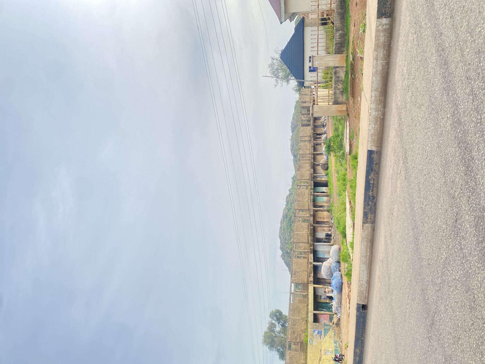 The image shows a street view in Jos, Nigeria, with a focus on a row of unfinished or dilapidated storefronts in the mid-ground. The storefronts appear to be made of concrete and have open entrances, some with visible debris and materials inside. In front of the storefronts, there are large, rounded objects covered in blue tarps, possibly bales of goods or waste. A motorcycle is parked to the left of the storefronts, and a person can be seen near the blue-covered objects. Behind the storefronts, a hilly landscape with green vegetation is visible, with some rocky outcrops. To the right of the storefronts, a modern house with a dark, gabled roof and a metal fence is present. The street in the foreground is paved and has a concrete curb with black and white markings. The sky is overcast with some lighter areas indicating sunlight. Power lines are stretched across the upper portion of the image. The overall scene depicts a developing or somewhat neglected urban area.
