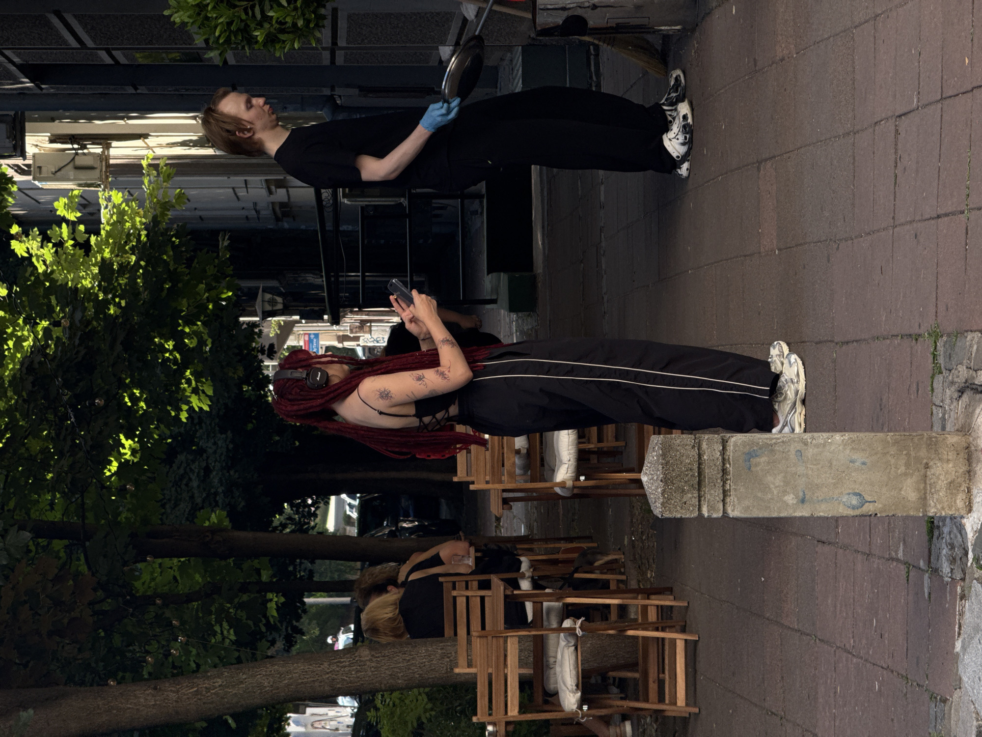 The image depicts an outdoor scene on a sunlit sidewalk in Belgrade, Serbia, likely near a cafe or restaurant, given the presence of outdoor seating. In the foreground, a person with distinctive red dreadlocks, wearing a black tank top and black pants with white stripes, is standing and looking at their phone. They are also wearing headphones. To the right, a person in a black t-shirt and black pants is holding a frying pan and wearing a blue glove. They appear to be interacting with a bin and a broom. In the background, several empty wooden chairs and tables are visible, along with trees providing shade. The overall atmosphere is that of a casual urban environment during daylight.