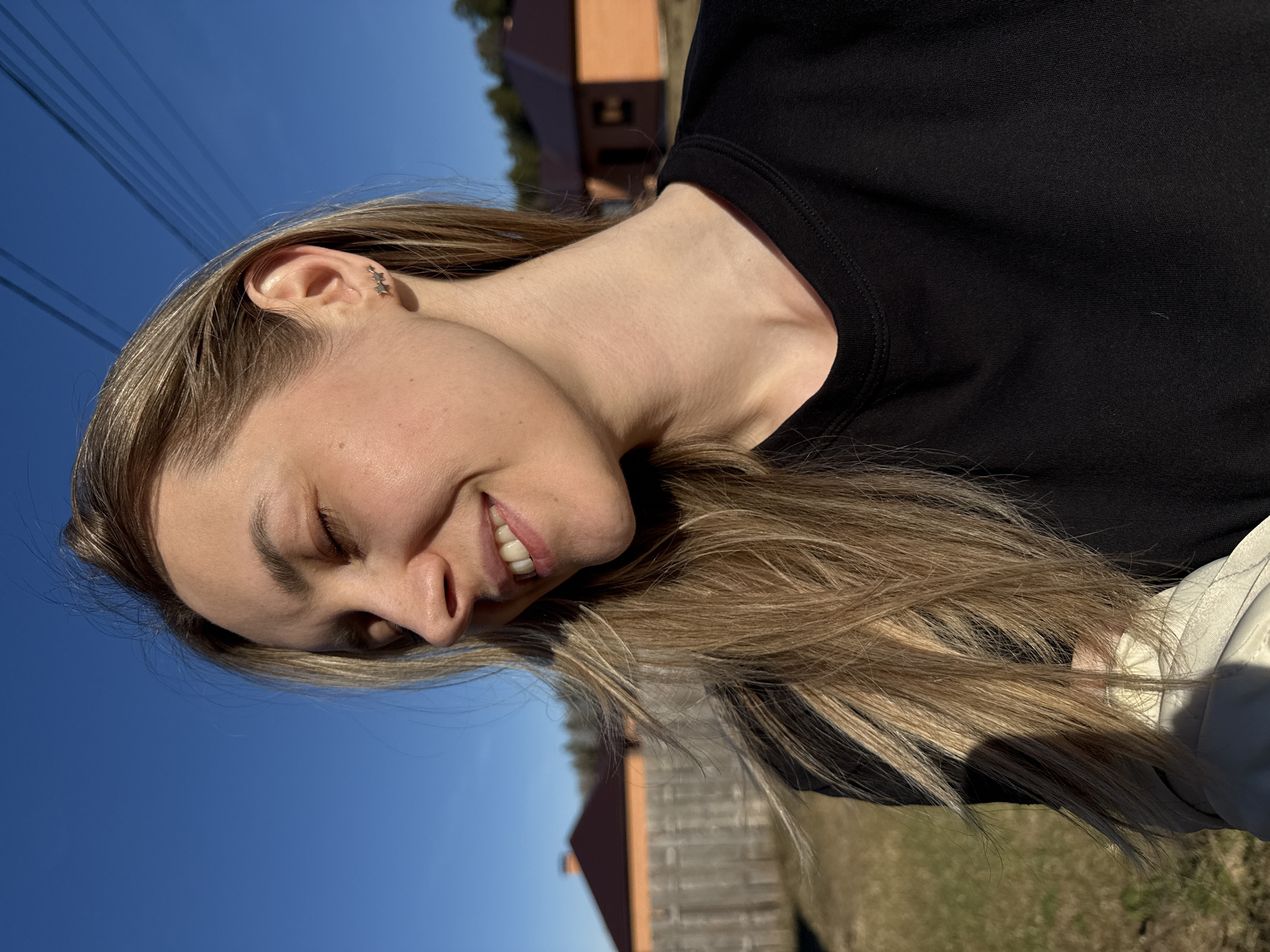 A young woman is captured in a vertically oriented portrait, rotated 90 degrees counter-clockwise. Her head, positioned on the left side of the frame, is tilted with her face angled upwards towards a bright light source. She has closed or squinting eyes, suggesting she is looking into the sun, and a broad, joyful smile, revealing her teeth. Her long, light brown or blonde hair cascades around her head and over her right shoulder. She wears a plain black top, visible on her right side. A small, distinctive silver earring, shaped like multiple stars, is visible in her left ear.
The background is dominated by a clear, vibrant blue sky. Several dark utility lines stretch horizontally across the upper part of the frame, against the sky. In the mid-ground, blurred structures, possibly houses or buildings with reddish-brown roofs and walls, are partially visible. The ground below these structures appears grassy or covered in dry foliage. The strong, direct sunlight indicates a sunny day, likely mid-day or afternoon, creating bright highlights on her hair and face. The scene conveys a relaxed and happy emotion in an outdoor setting in Chaikovsky, Russia. No visible text is present.