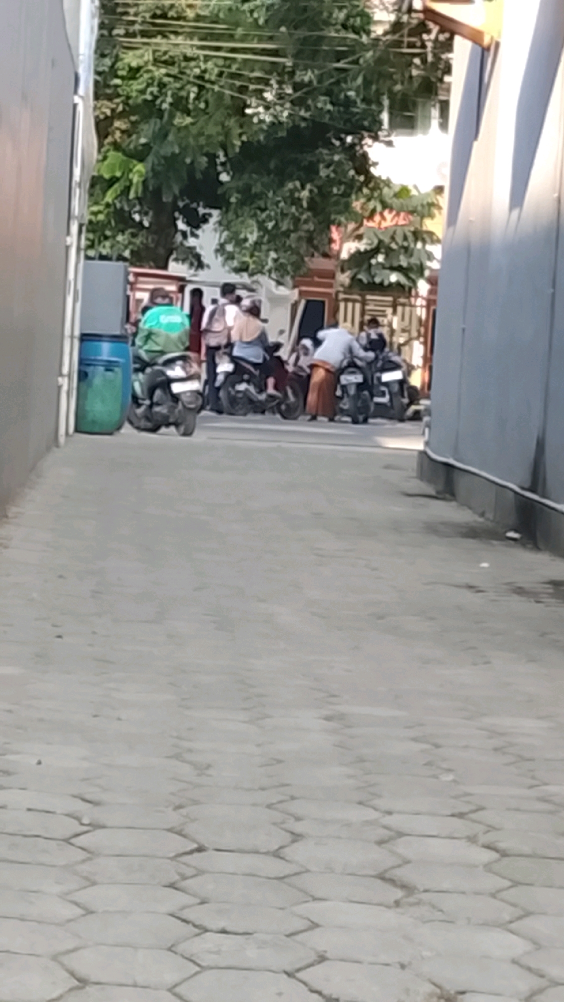 In Slawi, Indonesia, a group of people are gathered on a paved pathway lined with hexagonal bricks. On the left, a green trash bin stands next to a building. Several motorcycles are parked and occupied by individuals. One person, wearing a green jacket with the "Grab" logo, sits on a motorcycle. Others are on or near their motorcycles, with some appearing to be preparing to leave. The background shows trees and buildings. The scene is illuminated by sunlight, casting shadows.