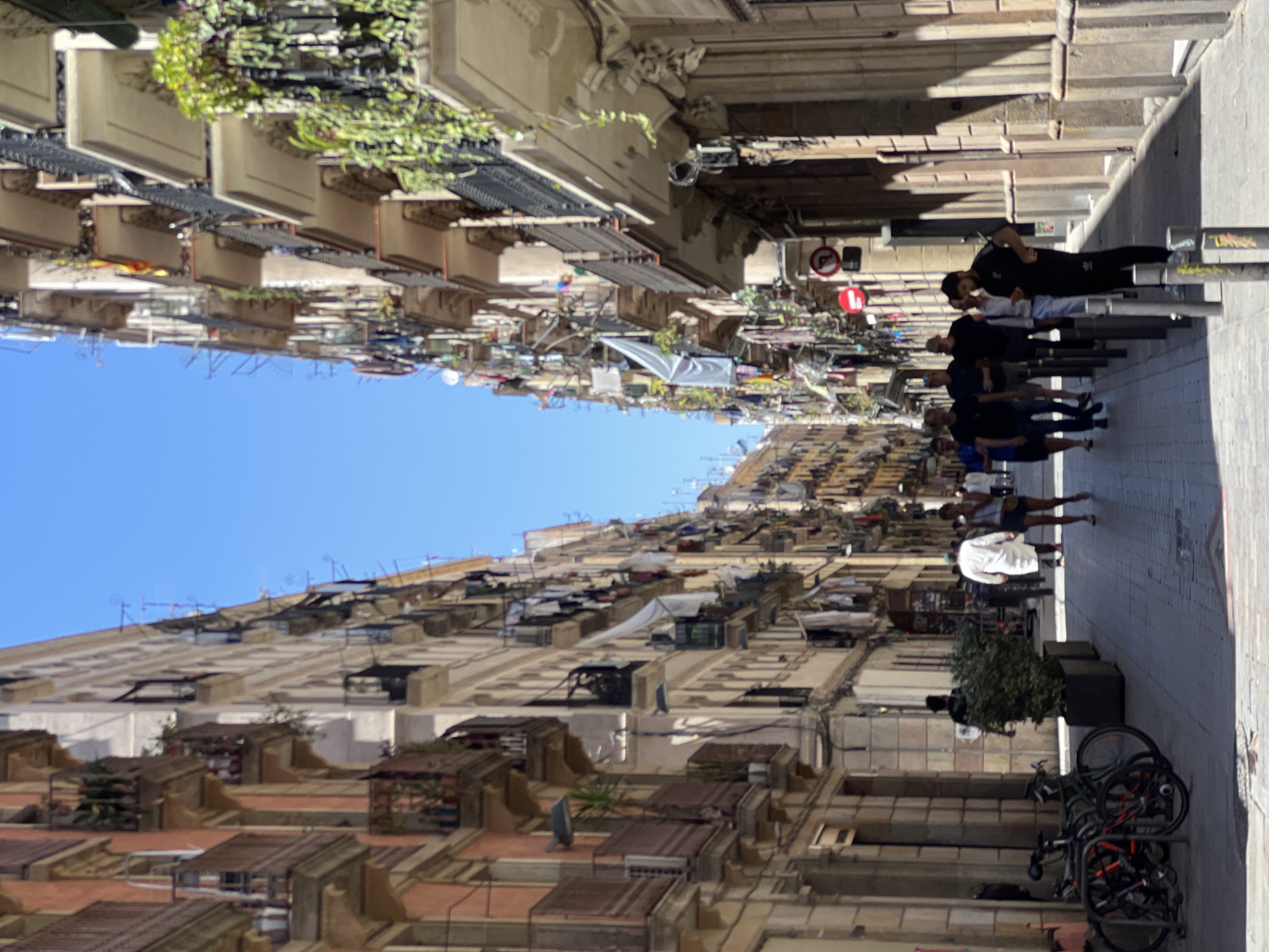 The image shows a narrow street in Barcelona, Spain, lined with tall, multi-story apartment buildings. The buildings have many balconies adorned with plants and laundry hanging out to dry. A few people are walking along the street, which is paved with cobblestones. There are bicycles parked on the left side of the street. The sky is clear and blue. The architecture suggests a historic European city.