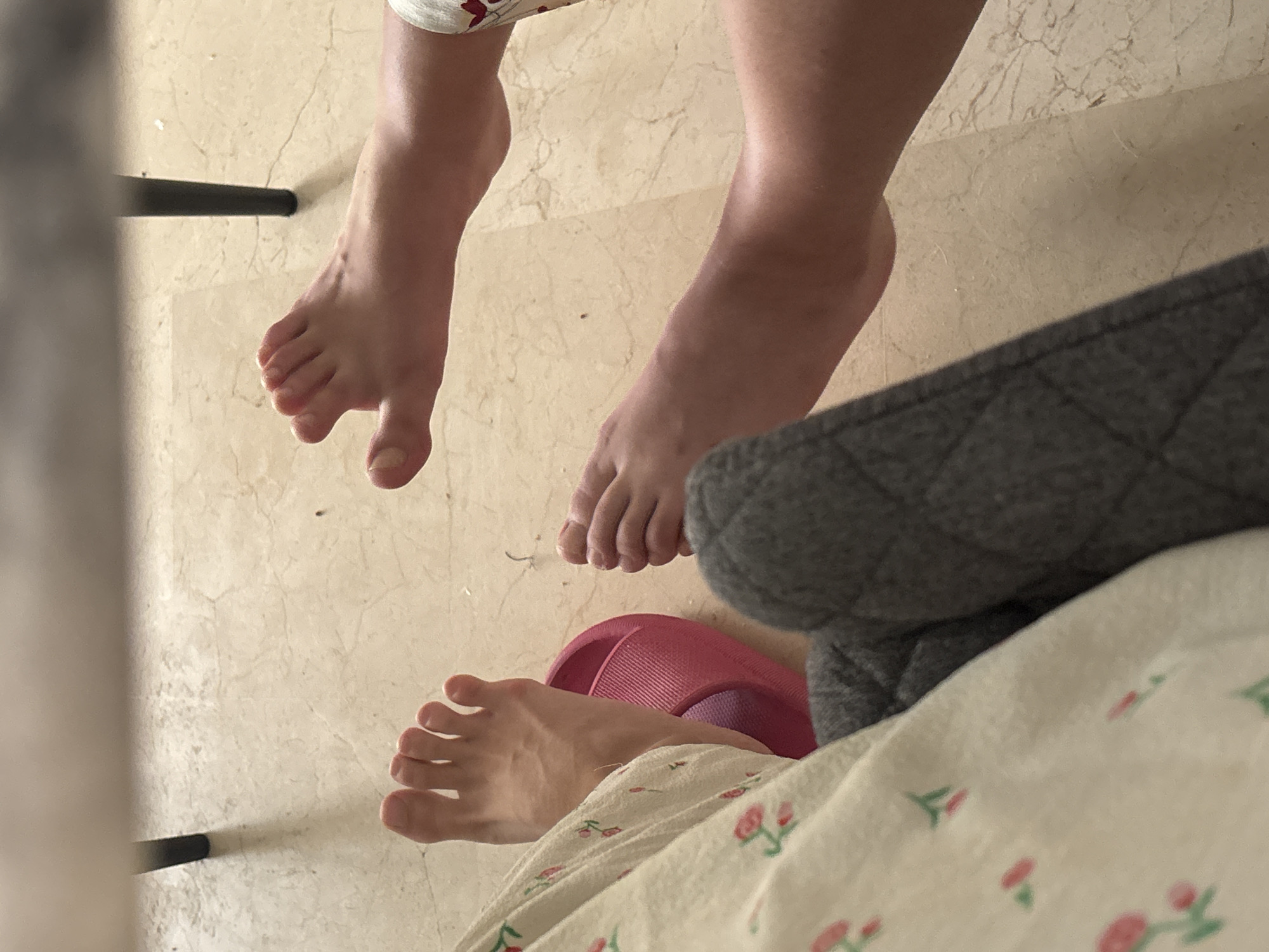 The image shows a close-up view of two people's feet resting on a tiled floor. One person's feet are visible on the left side of the frame, wearing a pink slipper. The person is also partially covered by a patterned fabric, possibly clothing or a blanket. The other person's feet are on the right side of the frame, and their legs are visible from the knee down, bare. Both individuals appear relaxed, with their feet positioned naturally on the floor. The floor is a light-colored tile with subtle variations in color and texture, suggesting it is likely indoors. The background includes the leg of a dark-colored table or chair. The image is shot from a high angle, looking down at the feet. There is no indication of the specific location within Orihuela, Spain.
