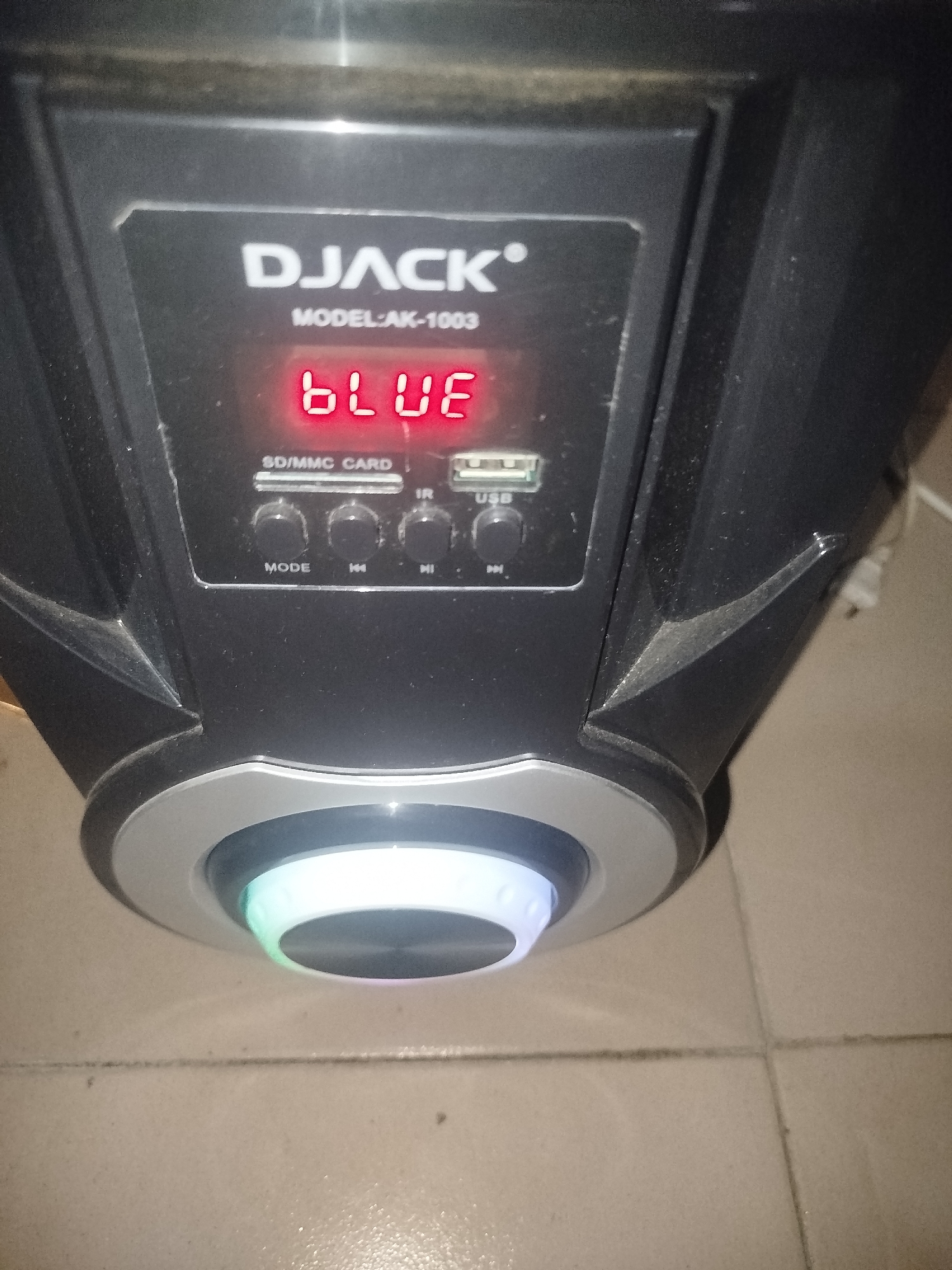 An image shows a DJACK AK-1003 model speaker. The speaker has a digital display showing "BLUE" in red LED lights. Below the display are buttons labeled "MODE", "<<", ">>", and "MI", along with indicators for "SD/MMC CARD", "IR", and "USB". At the bottom of the speaker, a ring light is illuminated with a gradient of blue and green colors. The speaker is positioned on a tiled floor. The scene is indoors in an unknown location.