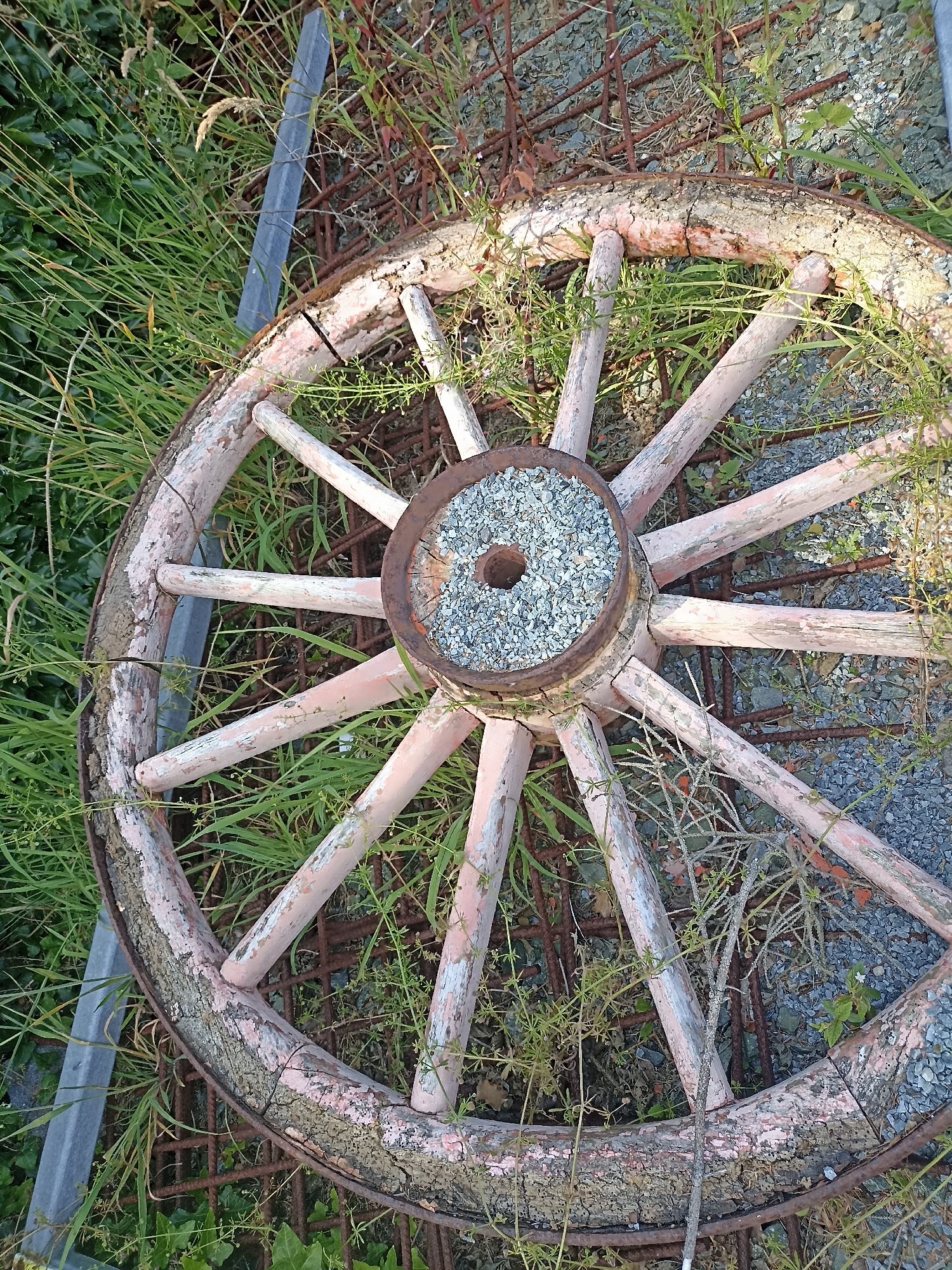 An old, weathered wagon wheel lies horizontally on the ground, partially covered by grass and weeds. The wheel is made of wood with spokes radiating from a central hub filled with small stones and gravel. The wooden rim is cracked and peeling, showing signs of age and decay. A rusty metal band encircles the outer edge of the wheel. The background consists of gravel, more overgrown vegetation, and some rusted metal bars. The scene suggests a rural or neglected outdoor setting. The image does not contain any people or depict any actions or interactions. The location in Doirí Beaga, Ireland is not discernible from the visual information in the image itself.