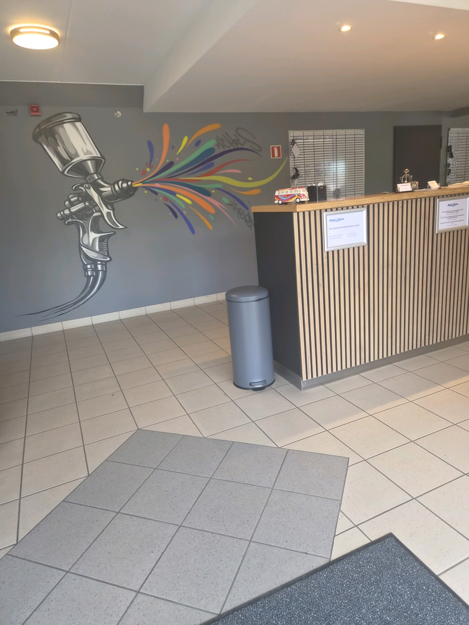 The image captures the interior of what appears to be a reception area or service counter in Sanem, Luxembourg. Dominating the left wall is a large, stylized mural of a spray gun emitting a vibrant rainbow of colors. Adjacent to the mural, a red fire alarm is mounted on the grey wall. In the center of the scene, a modern reception desk with vertical wooden slats is visible, topped with various items including a small toy bus, some documents, and desk accessories. On the wall behind the desk, a whiteboard or grid-like planner is displayed, and further to the right, a dark doorway is present. A grey, cylindrical trash can with a foot pedal stands to the left of the reception desk. The floor is tiled with a pattern of beige squares and a section of grey squares near the bottom of the frame. The lighting in the room comes from ceiling-mounted fixtures.