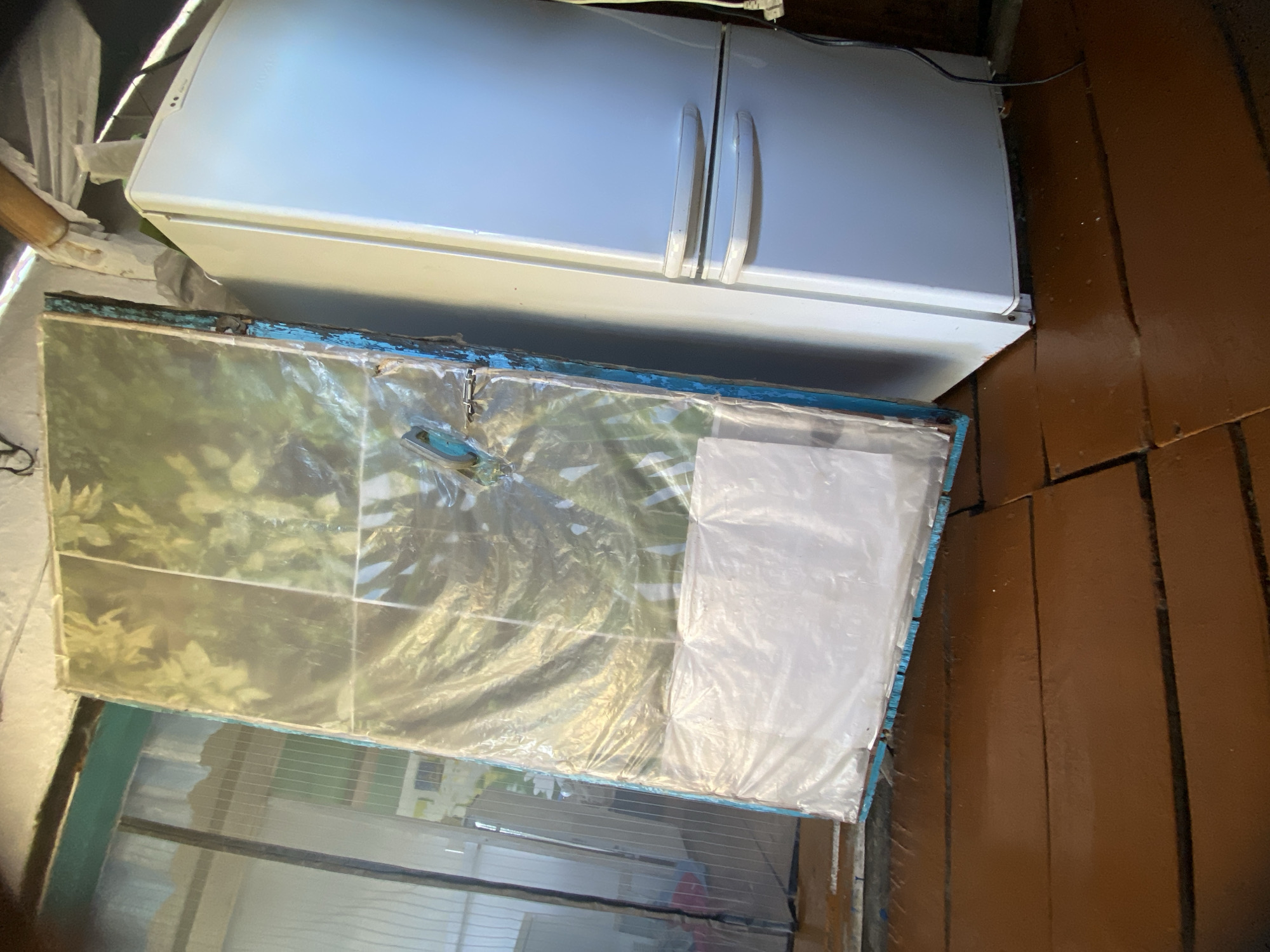 In a room with wooden flooring and a white ceiling, a white refrigerator stands to the right. To the left of the refrigerator, leaning against an unseen surface, is a large, rectangular object wrapped in clear plastic. This object has a frame that appears to be painted blue with some chipped paint. The plastic wrapping displays a pattern of green leaves. The overall scene suggests a storage or makeshift indoor area, possibly in a residential setting in Maloarslangulovo, Russia.
