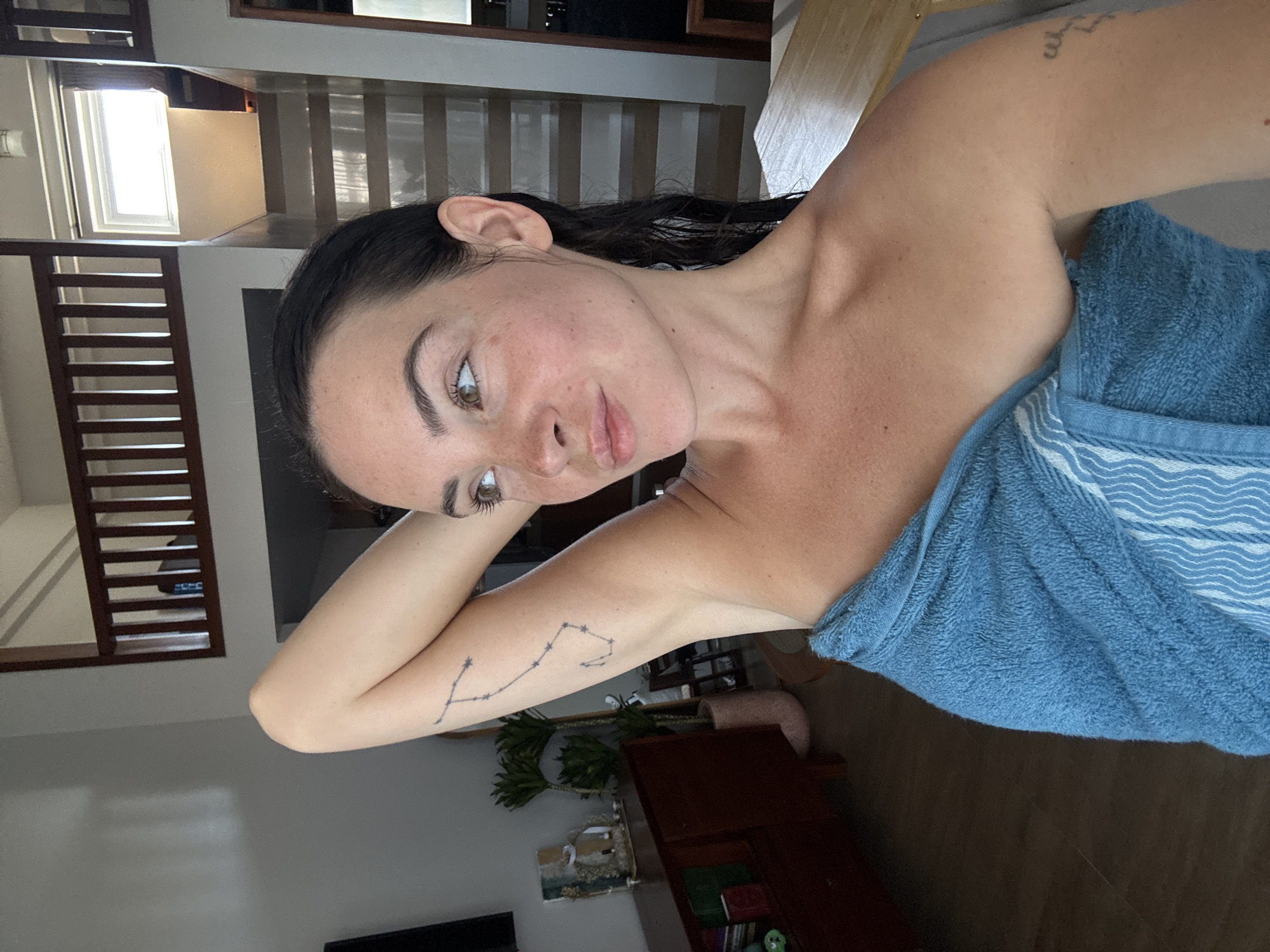 A woman with dark hair, wrapped in a blue towel, is pictured in an indoor setting that appears to be a residence in North Kuta, Indonesia. She is looking to her right, with her left arm raised and bent at the elbow, resting on her head. A tattoo of a constellation is visible on her upper left arm. In the background, a wooden railing and staircase suggest a lofted area within the house. A wooden cabinet with decorative items and plants is visible to the left of the woman, and a wooden table is in the foreground to her right. The lighting suggests it is daytime.
