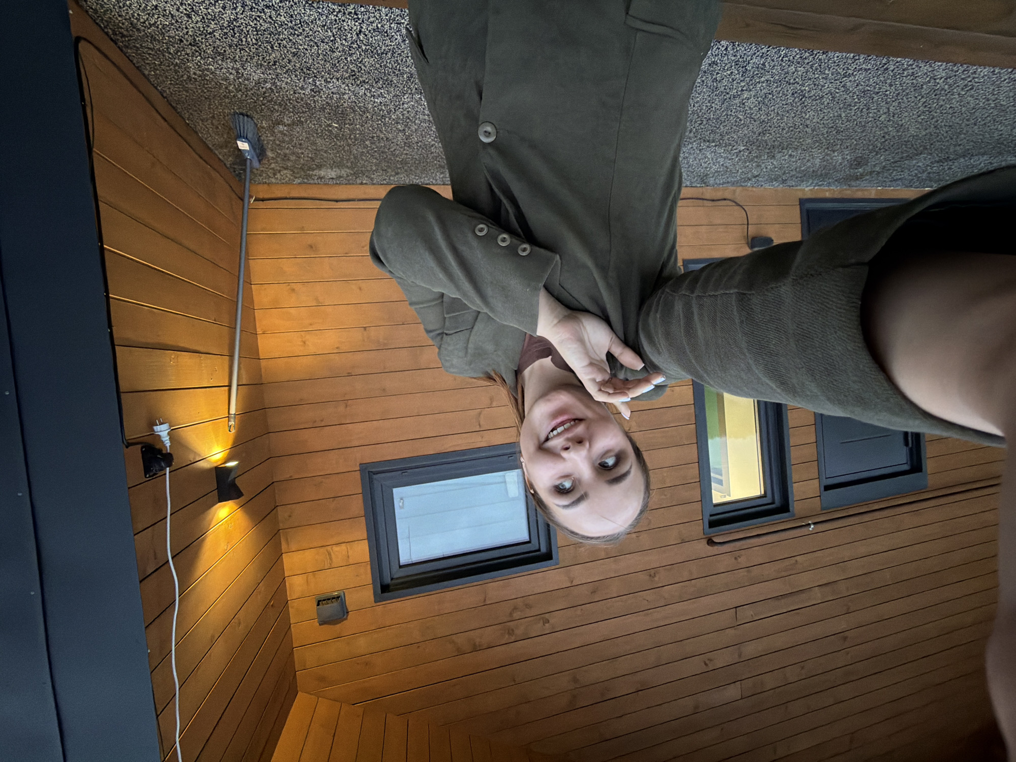 A young woman is taking a selfie outdoors in front of a wooden house. She is wearing a dark green blazer over a brown shirt. Her hair is pulled back in a ponytail. The house has dark trim around the windows and doors. There is a light fixture mounted on the wall and a broom leaning against the side of the house. The ground is covered in a grey carpet-like material. The woman is smiling and looking up and to her right. The image appears to be taken during the evening, as indicated by the artificial lighting. There is no information in the image to suggest this location is in Kamyennoye, Russia.