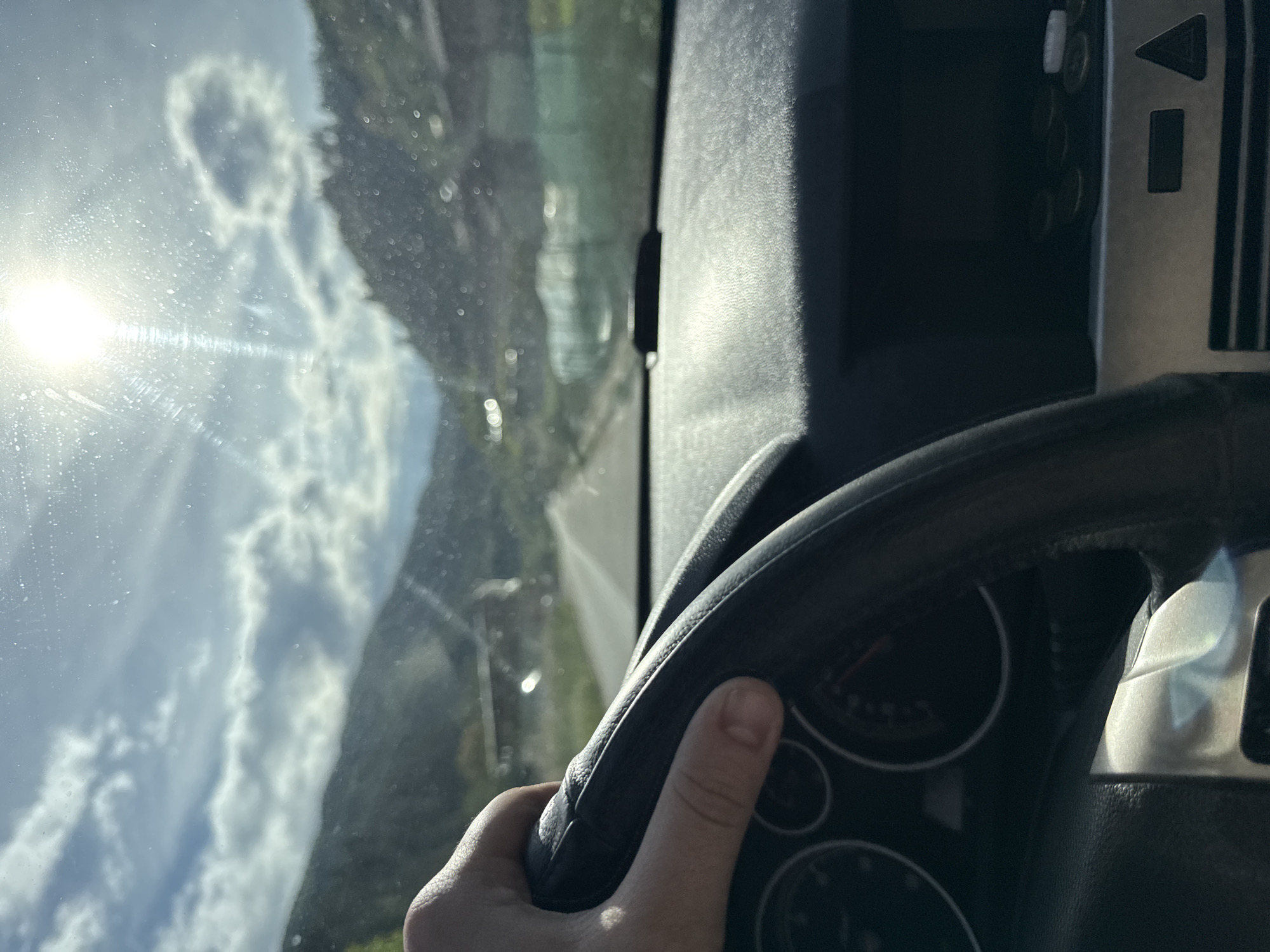 A driver's hand grips a black steering wheel inside a vehicle. Behind the wheel, through the windshield, is a landscape of tree-covered mountains and a partly cloudy sky, the sun a bright spot in the upper left corner. Based on the geography visible, it is highly probable that the scene is from Byala Reka, Bulgaria, given the mountainous terrain. The vehicle's dashboard and controls are visible to the right of the steering wheel.