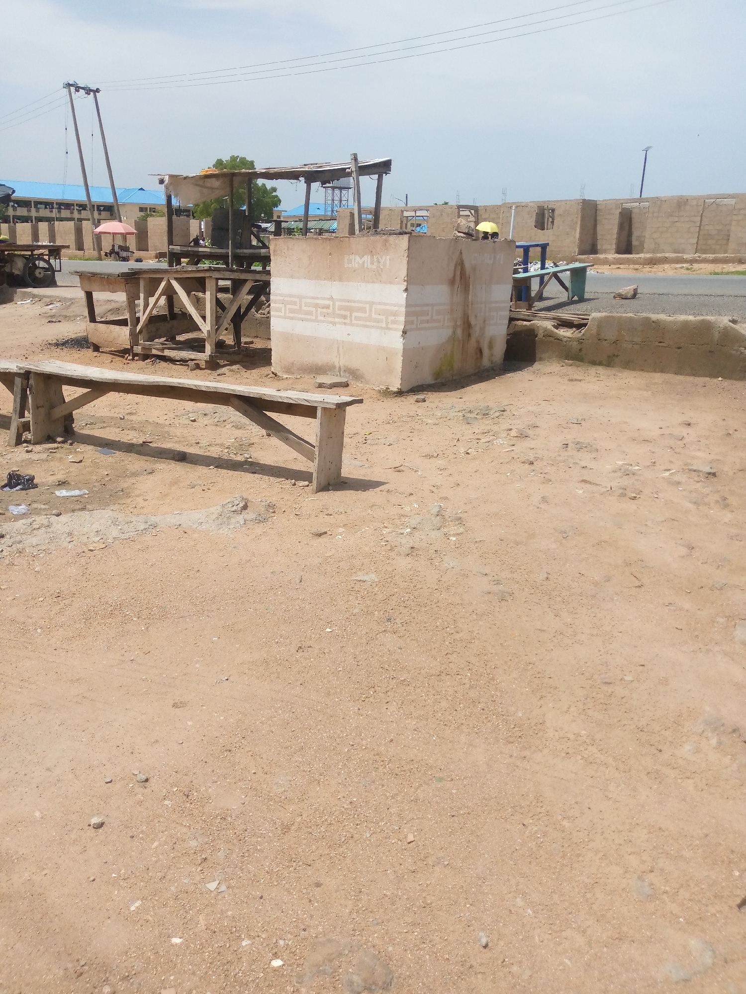 The image depicts an outdoor scene in Jalingo, Nigeria, likely a marketplace or construction site. In the foreground, a long wooden bench sits on dusty, uneven ground. Behind it, a rough, cubical concrete structure with decorative markings is visible. To the left, a makeshift wooden stall with a roof of corrugated metal and fabric stands. Further in the background, a line of unfinished concrete buildings suggests ongoing construction. An umbrella, a cart with wheels, and a blue table with a green bench are also present in the middle ground. Power lines and poles crisscross the sky. The overall impression is one of a developing area with a mix of temporary structures and new construction under a bright, overcast sky.