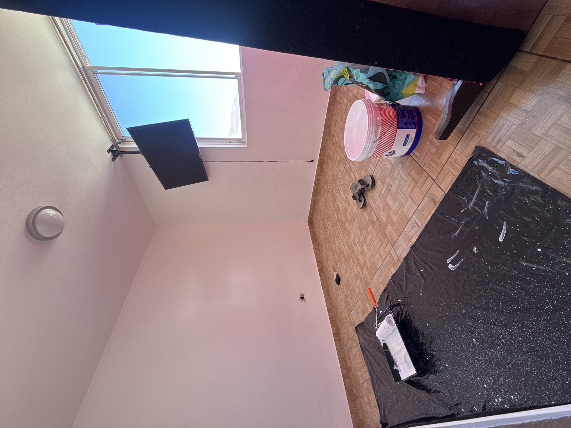 The image depicts an interior scene, likely a room undergoing renovation, located in the city of Iquique, Chile. The walls are painted a light pink, and the floor is tiled in a brown, square pattern.
In the foreground, a black tarp is spread on the floor, covered with white paint splatters and streaks. A paint roller with an orange handle rests in a black tray on the tarp, containing white paint. To the right of the tarp, a white bucket with red and blue labeling is visible, containing white paint. Next to the bucket are a pair of dark flip-flops and a partially obscured object, possibly a shopping bag with a colorful pattern.
A flat-screen television is mounted on the wall near a large window, which offers a view of a clear blue sky and a distant landscape. The window has a white frame. A ceiling light fixture is present in the upper left corner of the room. In the background, against the far wall, a dark, upright object, possibly furniture, is partially visible.