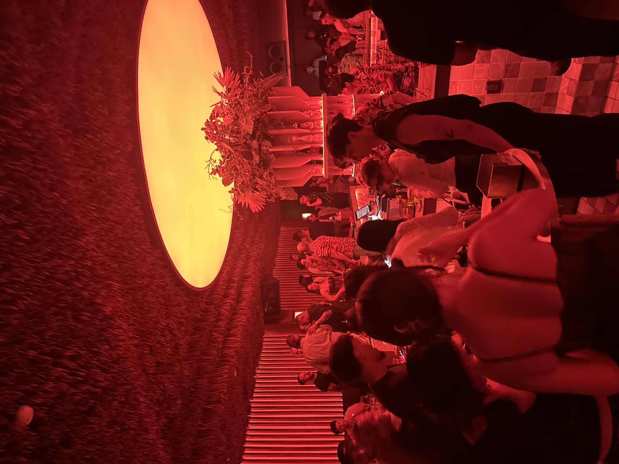 A dimly lit, red-toned bar in Thủ Đức, Vietnam, is crowded with people. A large, circular light fixture dominates the left side of the image, partially obscured by decorative foliage. A bar runs horizontally across the frame, with several individuals standing or sitting along it. Bottles are visible on shelves behind the bar. People are interacting in small groups, engaging in conversation and drinking. The floor is tiled, and steps lead down into the bar area. The image is rotated counter-clockwise.