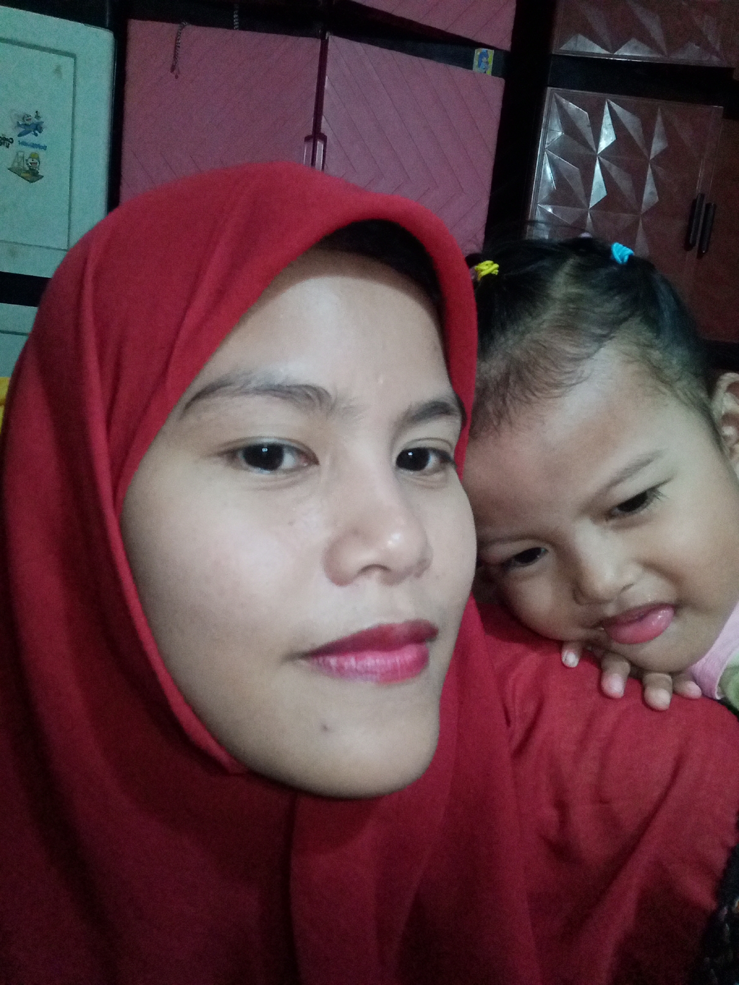 A close-up, waist-up selfie shows a woman in a red hijab and a young child. The woman's face is turned towards the camera, with her left eye and lips visible. She is wearing bright pink lipstick. The child is partially visible behind the woman, resting their head on the woman's shoulder. The child's face is turned away from the camera, with their mouth slightly open and tongue sticking out. The child has a few colorful hair ties in their hair. The background is dark and appears to be a room with furniture. The lighting is artificial and casts shadows. The image was taken indoors, likely in a residential setting.