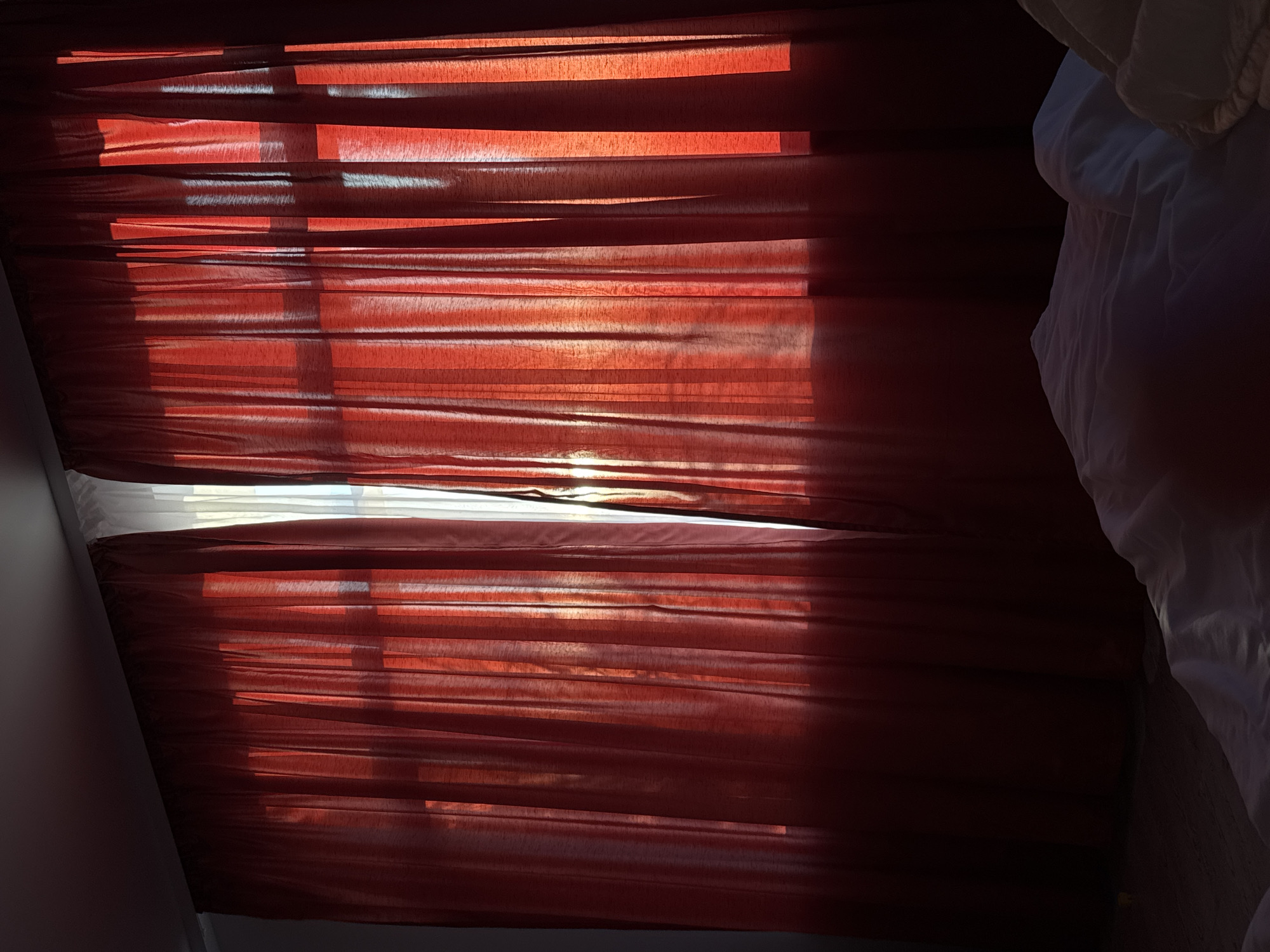 The image shows red curtains partially covering a window, with sunlight filtering through the gaps. Part of a white bed sheet is visible on the right. The exact location in Saint Petersburg, Russia is not discernible from the visual information provided.
