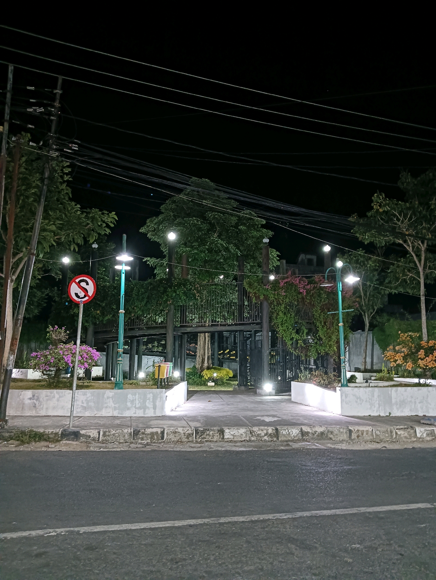 The image captures an outdoor scene at night, possibly in a public space within the city of Purworejo, Indonesia. The foreground features an asphalt road with a white line marking. Beyond the road is a concrete sidewalk and a low, white-painted retaining wall bordering a garden area.
In the garden, there are several trees and flowering bushes, some with pink and orange blooms. Streetlights, designed as decorative poles with multiple lamps, illuminate the area. A traffic sign with a red circle and a diagonal line over the letter "S" is visible on the left, likely indicating a "no stopping" or "no parking" regulation.
In the background, a multi-level structure, possibly a pavilion or elevated walkway, is visible amidst dense foliage. The structure appears to be made of metal and wood, with a dark fence or gate at its entrance. The night sky is dark, with visible power lines crisscrossing the upper portion of the image, supported by utility poles on the left. The lighting suggests the area is a well-maintained public space, potentially a park or a decorative roadside garden, illuminated for evening use. No people are visible in the image.