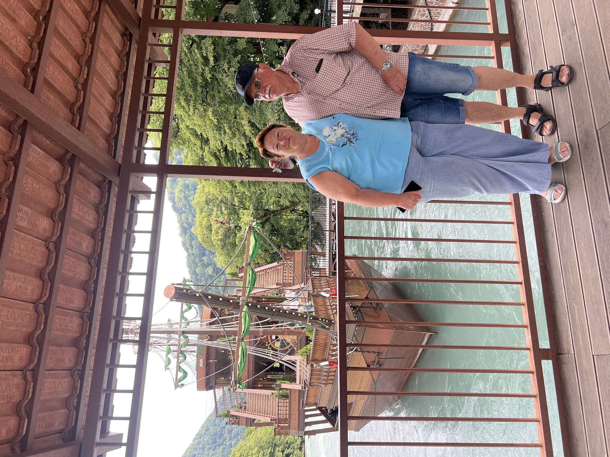 An outdoor medium shot captures a couple standing on a wooden deck with a railing in the foreground. Behind them, a large, ornate wooden pirate ship is docked on a turquoise river. The ship features multiple levels, masts with green decorative banners, and intricate wooden detailing. Lush green trees and mountains form the backdrop. The couple, a woman and a man, are positioned to the right of the frame, facing the camera. The woman wears a blue t-shirt and light blue pants, holding a black smartphone in her left hand. The man is dressed in a checkered shirt, denim shorts, and a blue baseball cap, with glasses on his nose and a watch on his left wrist. Both are wearing sandals. The scene appears to be a tourist attraction, possibly a restaurant or themed venue, in a scenic natural setting.