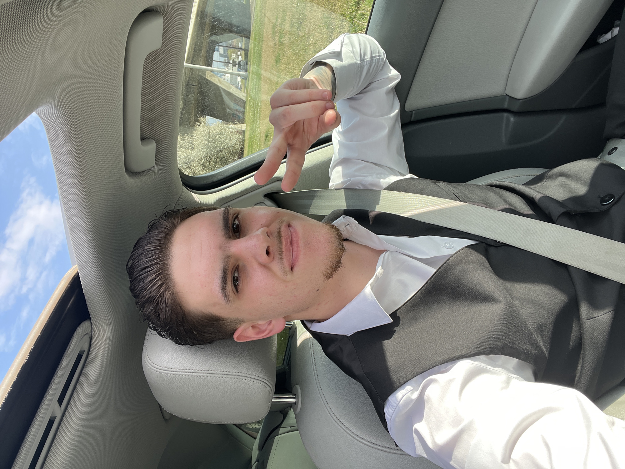 A young adult male, with dark, slicked-back hair, a mustache, and a short goatee, is seated in the front passenger position of a vehicle. He wears a white button-down shirt and a black vest, secured by a light grey seatbelt. His right hand is raised, forming a peace gesture. A light-toned tattoo is partially visible on his right forearm. The vehicle interior features light grey upholstery and a textured headliner with an open sunroof. Through the passenger window, a green grassy area is visible, alongside a multi-span concrete overpass and distant urban structures. The scene is illuminated by strong sunlight, indicating outdoor conditions in the city of Sofia, Bulgaria.