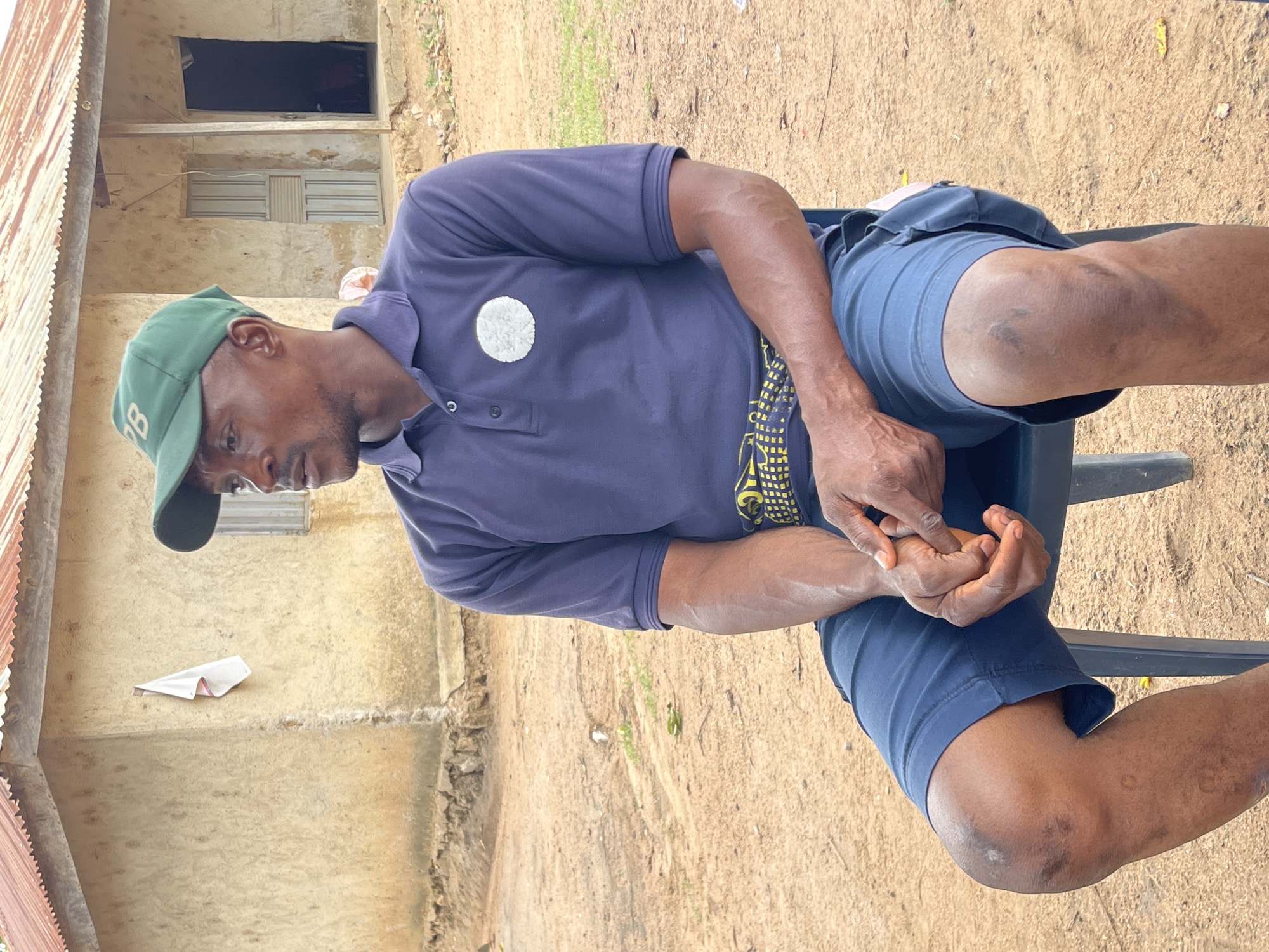 An outdoor, eye-level shot captures a man seated on a chair in a rural or semi-rural setting in Tabu, Nigeria. The man, with dark skin, is positioned in the foreground, looking downwards or to his left with a neutral expression. He wears a dark green baseball cap with a white letter "B" on the front, a dark blue short-sleeved polo shirt featuring a circular white, possibly textured, patch on the left chest, and dark blue shorts. His hands are clasped together in his lap, resting. Visible veins on his forearms and scuff marks on his knees suggest a history of physical activity or wear. A vibrant yellow and black patterned fabric is visible beneath his polo shirt, likely a wrapper or another layer of clothing.
The background consists of a simple, weathered building with light-colored, possibly painted, walls showing signs of age. An open, dark doorway and a window with horizontal metal slats are visible on the building. To the far left, a section of a corrugated metal roof is present. The ground beneath and around the man is dry, sandy earth. The scene is illuminated by bright daylight, indicating daytime and clear weather conditions. The overall atmosphere suggests an everyday moment in a rustic environment.